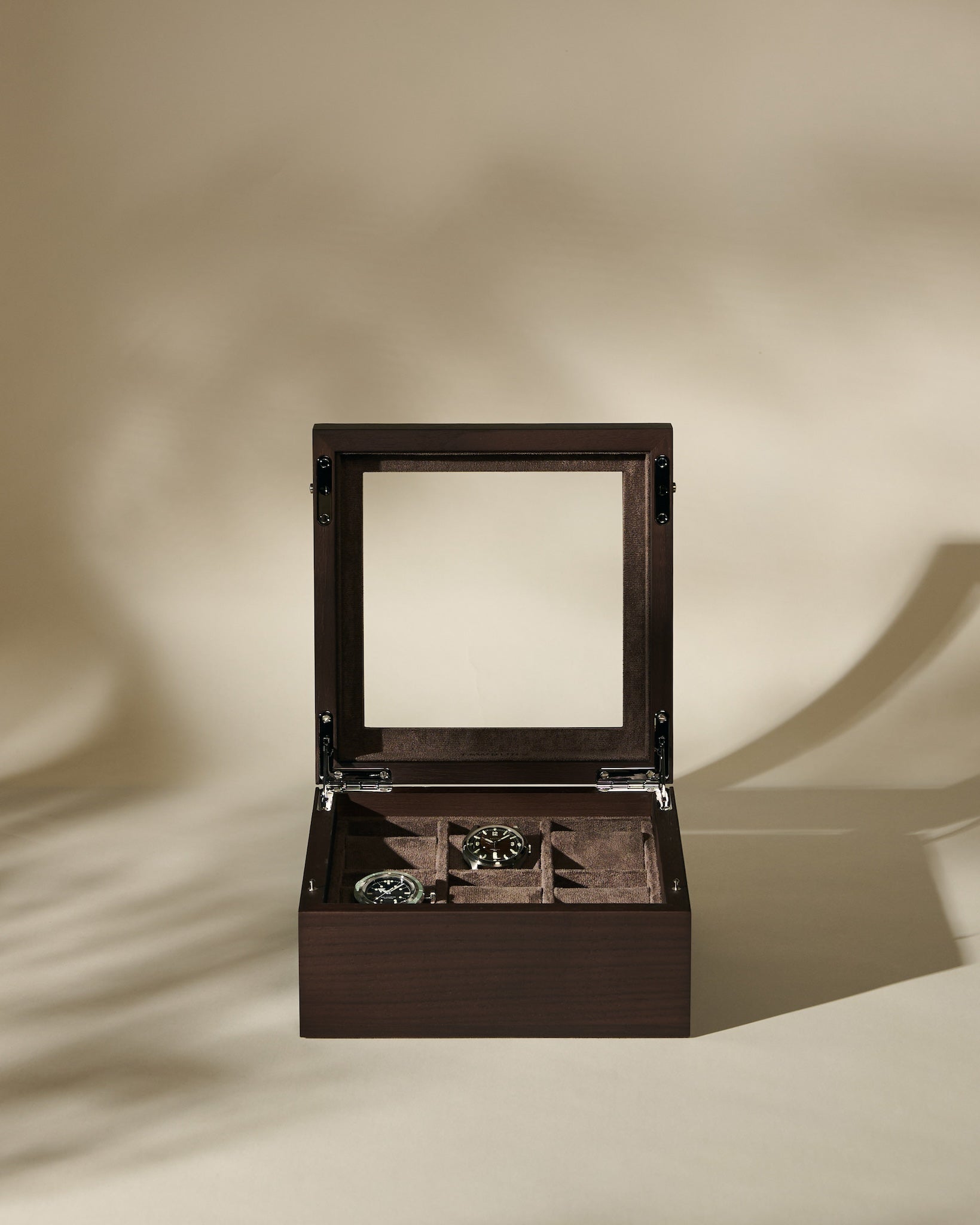A TAWBURY Grove 6 Slot Wooden Watch Box in Dark Walnut with a glass lid is open, stylishly presenting two wristwatches inside against a beige background.