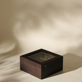 The TAWBURY Grove 6 Slot Wooden Watch Box in dark walnut wood features a glass lid, elegantly displaying a wristwatch inside on a neutral beige surface with soft shadows.
