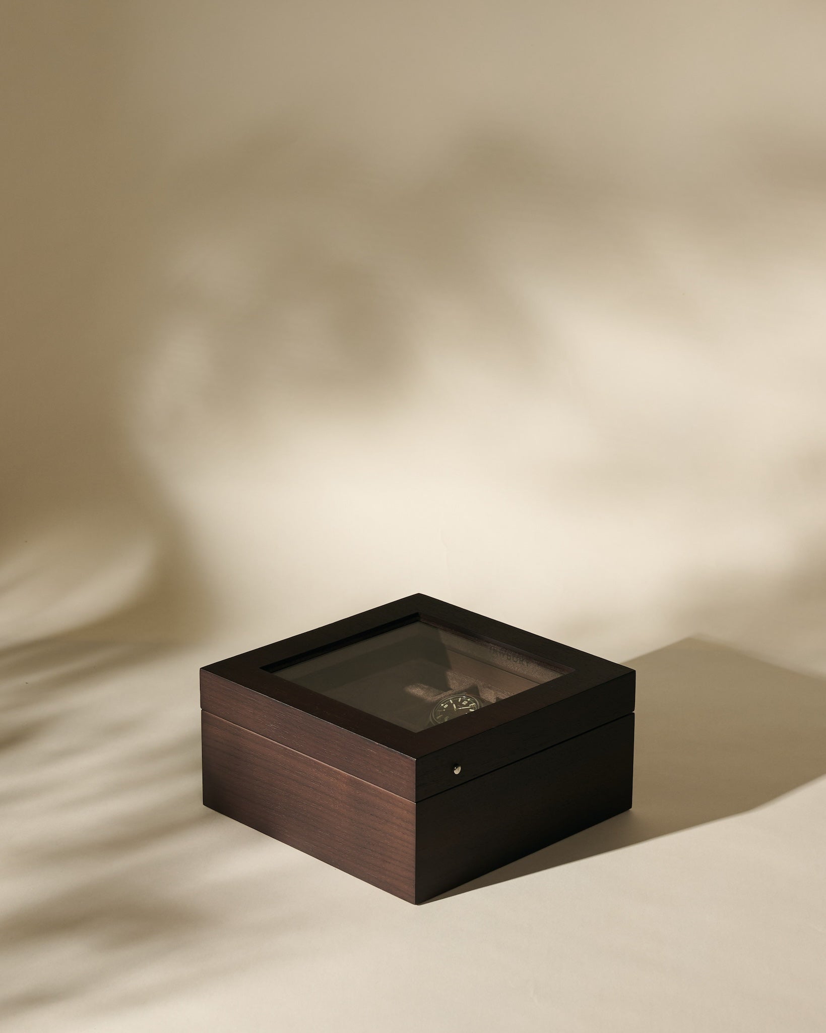 The TAWBURY Grove 6 Slot Wooden Watch Box in dark walnut wood features a glass lid, elegantly displaying a wristwatch inside on a neutral beige surface with soft shadows.