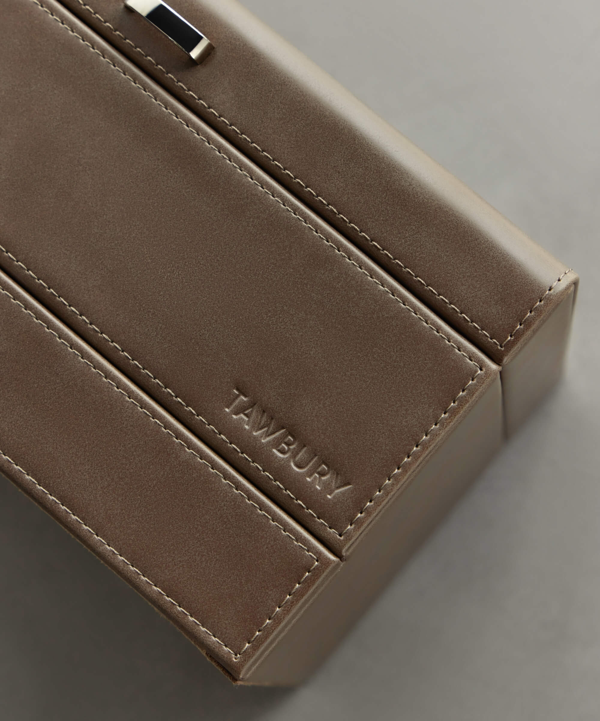 A close-up of the taupe Bayswater 12 Slot Watch Box with Storage by TAWBURY showcases its sleek vegan leather design, complete with clean lines and a silver clasp, set against a gray background—ideal for watch enthusiasts.