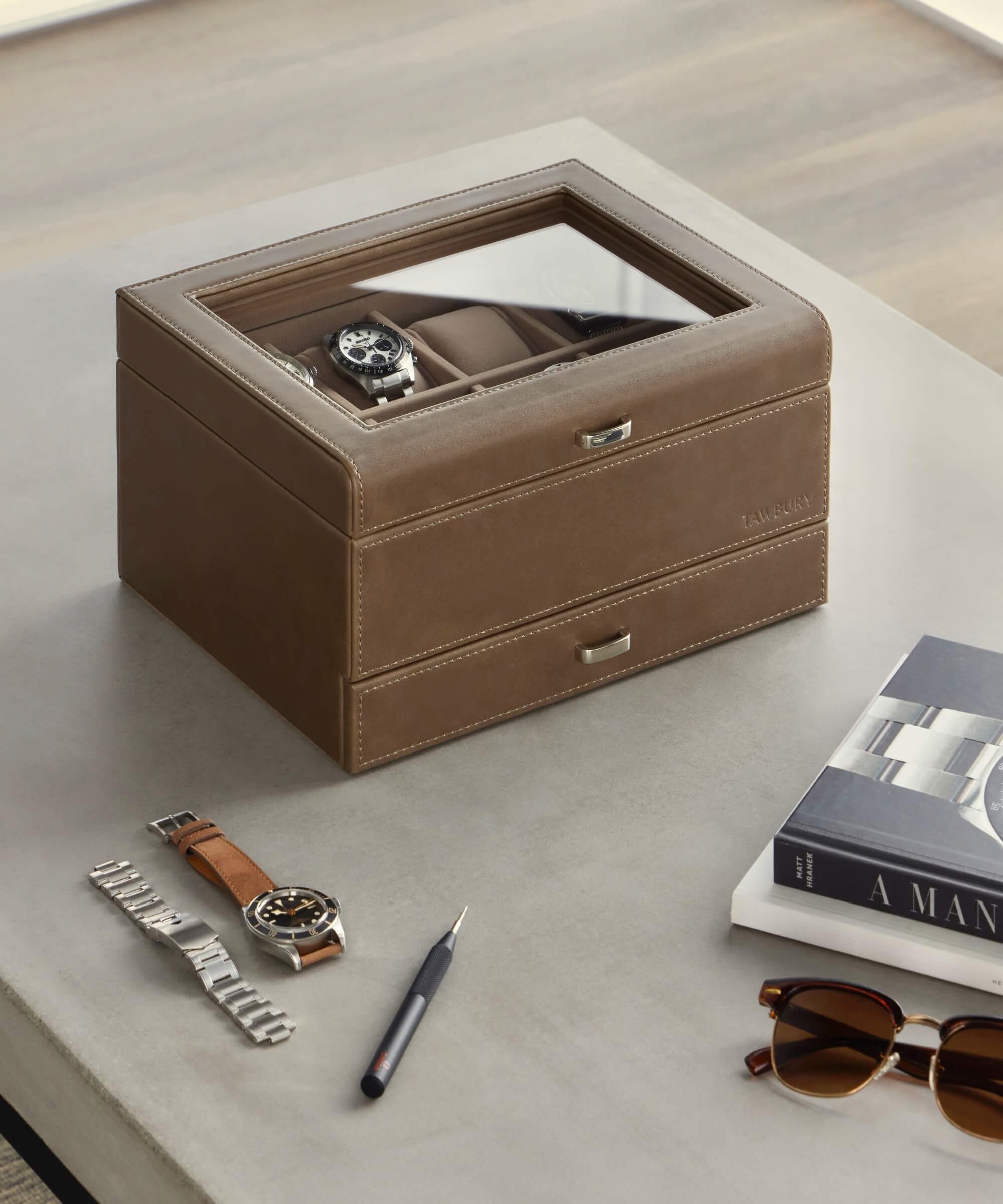 A taupe vegan leather watch box from TAWBURY’s Bayswater collection, featuring a glass top and designed to accommodate eight timepieces, adorns the table. Ideal for any watch enthusiast, this luxurious display is complemented by an elegant watch beside it, accompanied by a chic arrangement of a bracelet, pen, book, and sunglasses.