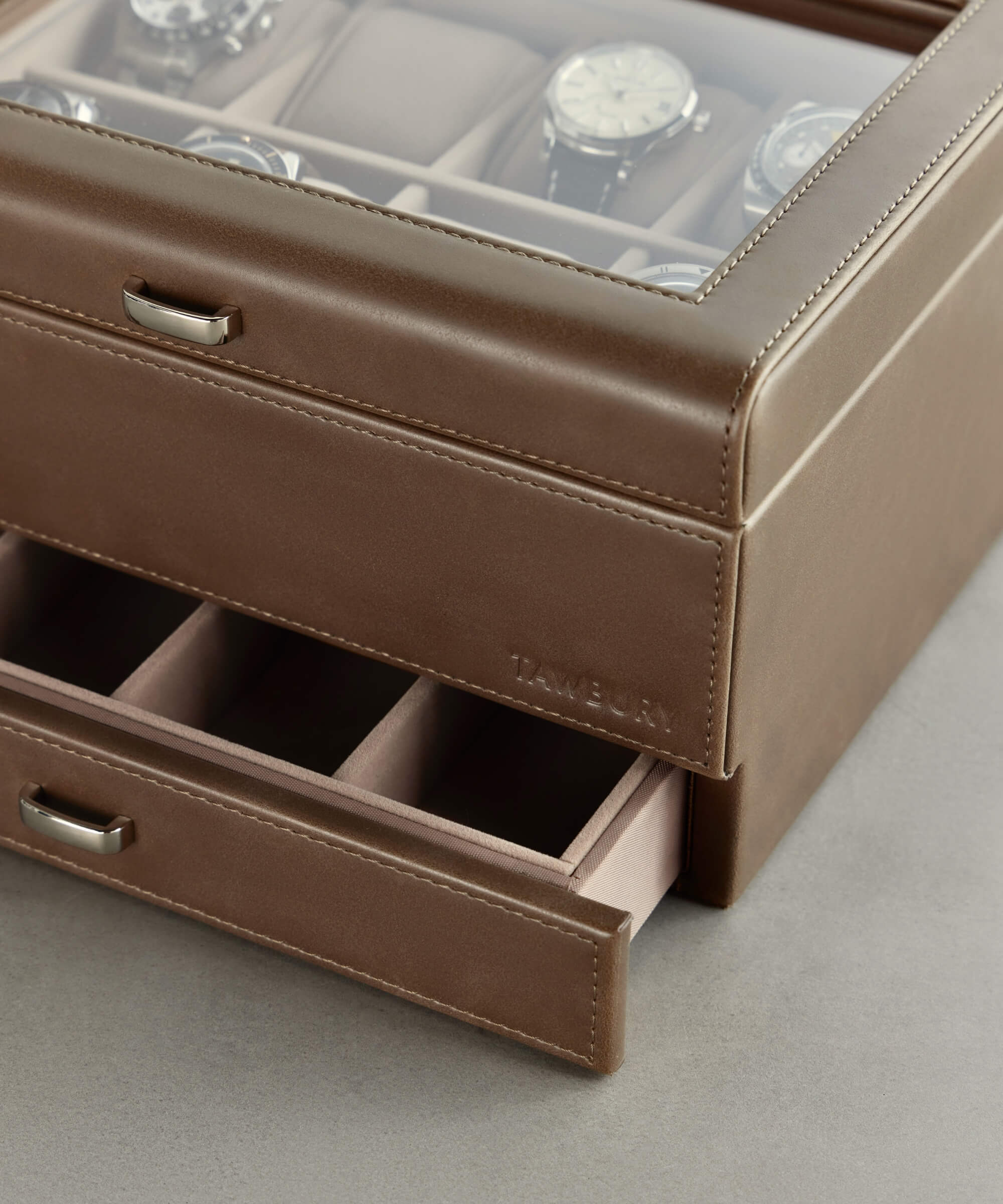 The Bayswater 8 Slot Watch Box with Storage by TAWBURY, crafted in luxurious taupe vegan leather, features a glass lid that elegantly displays your treasured timepieces. An open drawer below reveals additional compartments, making it perfect for any watch collector's essentials, offering both style and functionality.