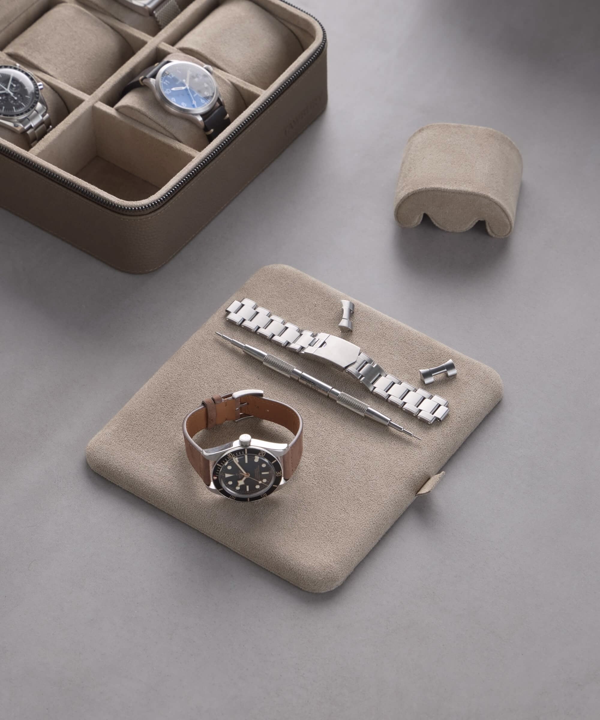 A brown watch with a metal bracelet and tools sits on a beige pad beside the TAWBURY Fraser 6 Watch Case in taupe, displaying exquisite timepieces.