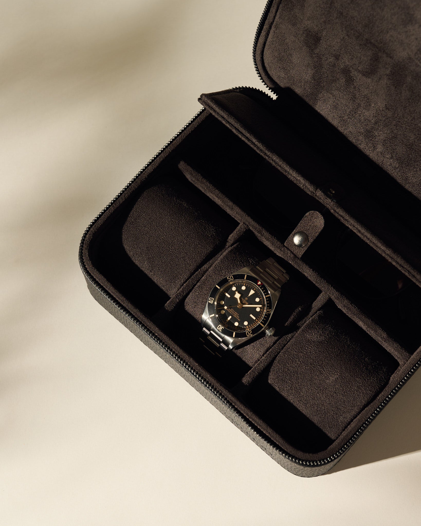 The TAWBURY Fraser 3 Watch Case with Storage - Black securely holds a wristwatch with a metallic band and black dial in its compartmentalized suede interior, displayed on a light surface.