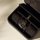 The TAWBURY Fraser 3 Watch Case with Storage - Black securely holds a wristwatch with a metallic band and black dial in its compartmentalized suede interior, displayed on a light surface.