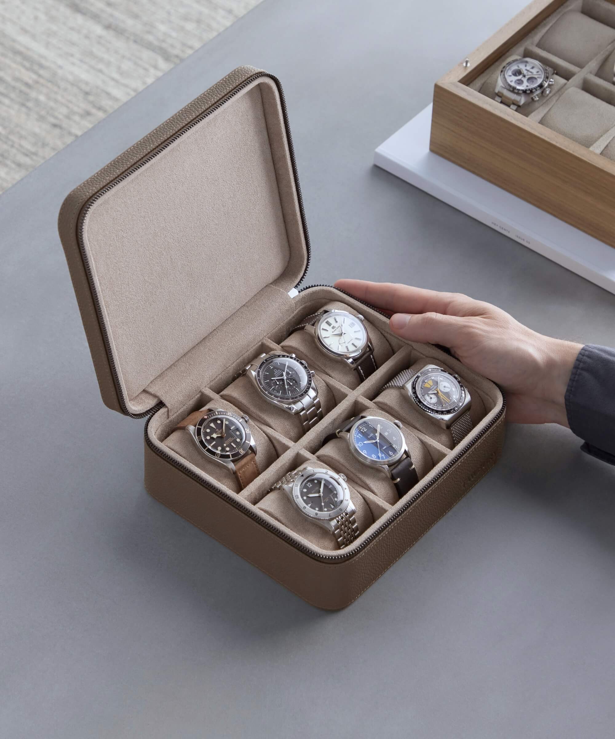 A person holds an open TAWBURY Fraser 6 Watch Travel Case - Taupe containing six wristwatches, with three more watches from the Fraser Collection visible in a separate box nearby.