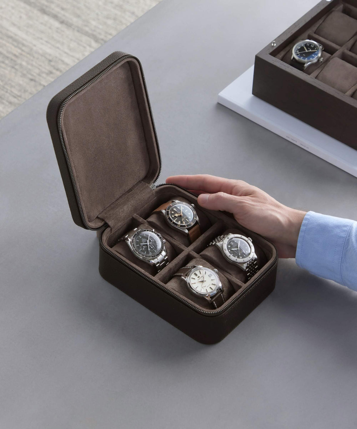 A hand is holding an open TAWBURY Fraser 4 Watch Travel Case in brown, containing four wristwatches. Another similar luxury leather watch case with wristwatches can be seen in the background.
