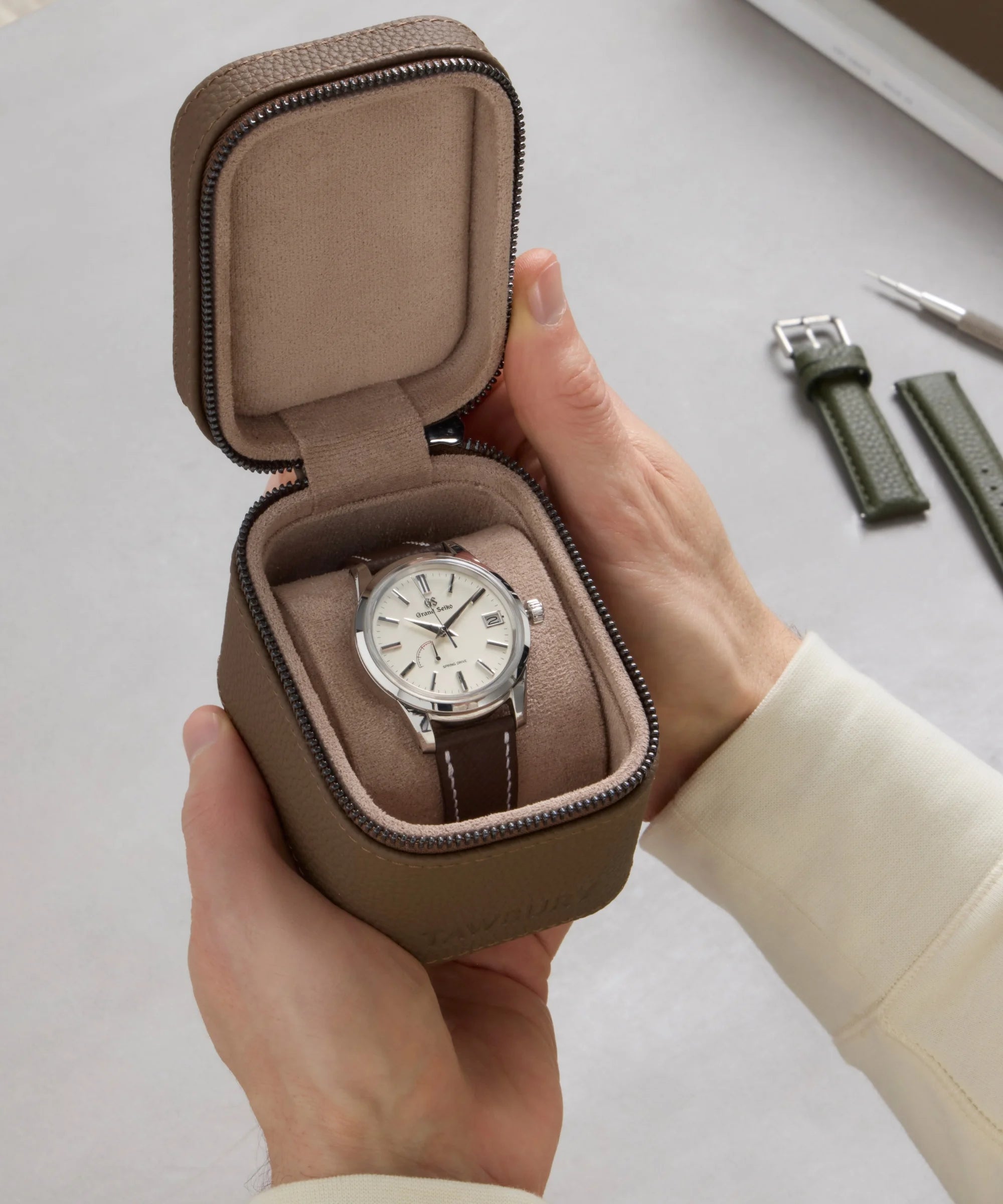 A person holds the TAWBURY Fraser 1 Watch Travel Case - Taupe (Coming Soon), displaying a wristwatch with a light face and brown strap, as extra straps and a tool are on the table.