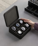 A person’s hand is holding an open Fraser 6 Watch Travel Case - Black (Coming Soon) from TAWBURY, showcasing six different wristwatches. Another closed luxury leather watch case from the TAWBURY range is in the background on a gray surface.