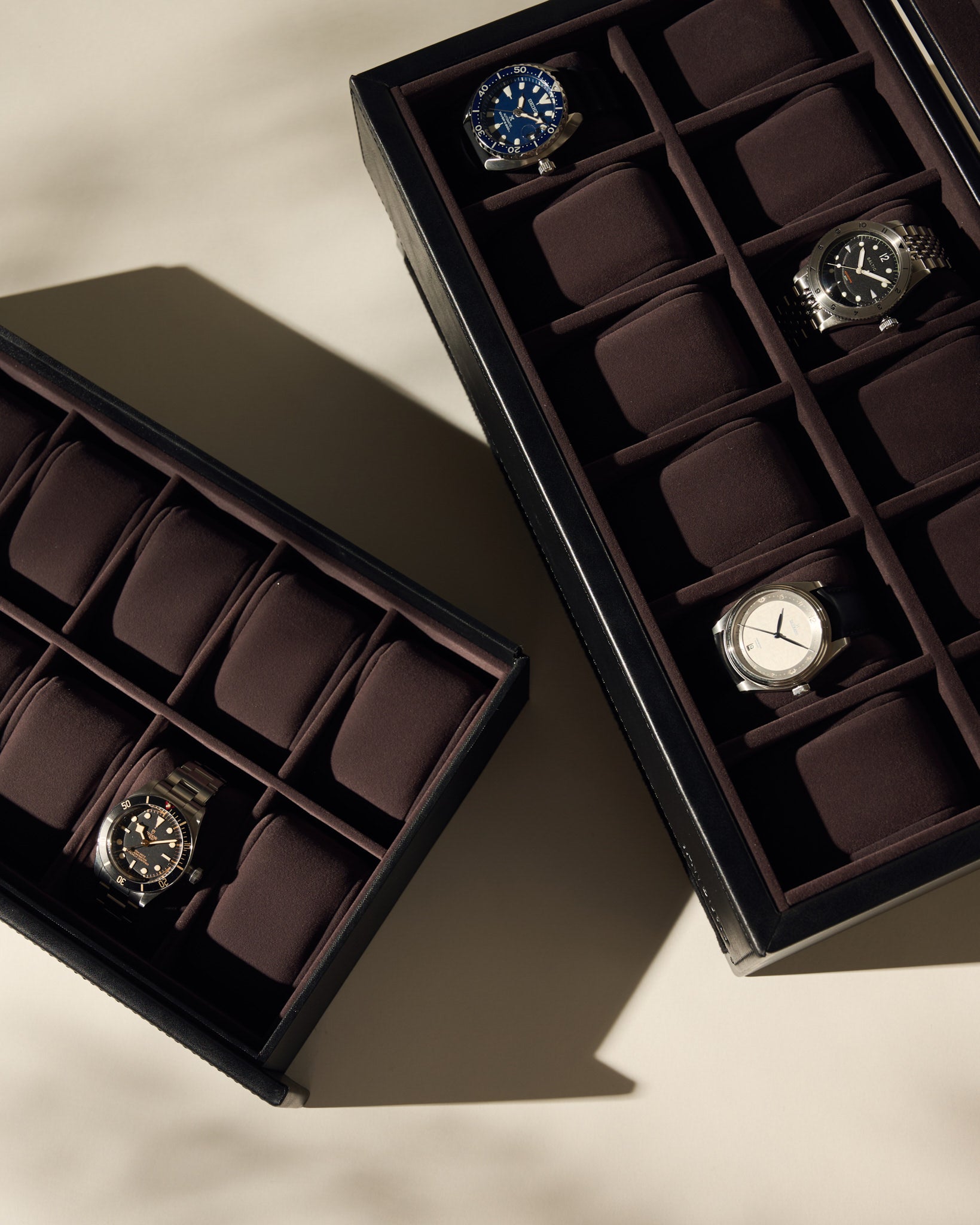 Two TAWBURY Bayswater 24 Slot Watch Boxes with Drawer in Black/Charcoal, each holding wristwatches with some empty slots visible, are displayed on a beige surface.