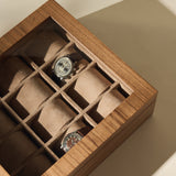 The TAWBURY Grove 12 Slot Wooden Watch Box in light walnut features a glass lid and displays two wristwatches on brown suede cushions, offering elegant storage while several slots remain empty.