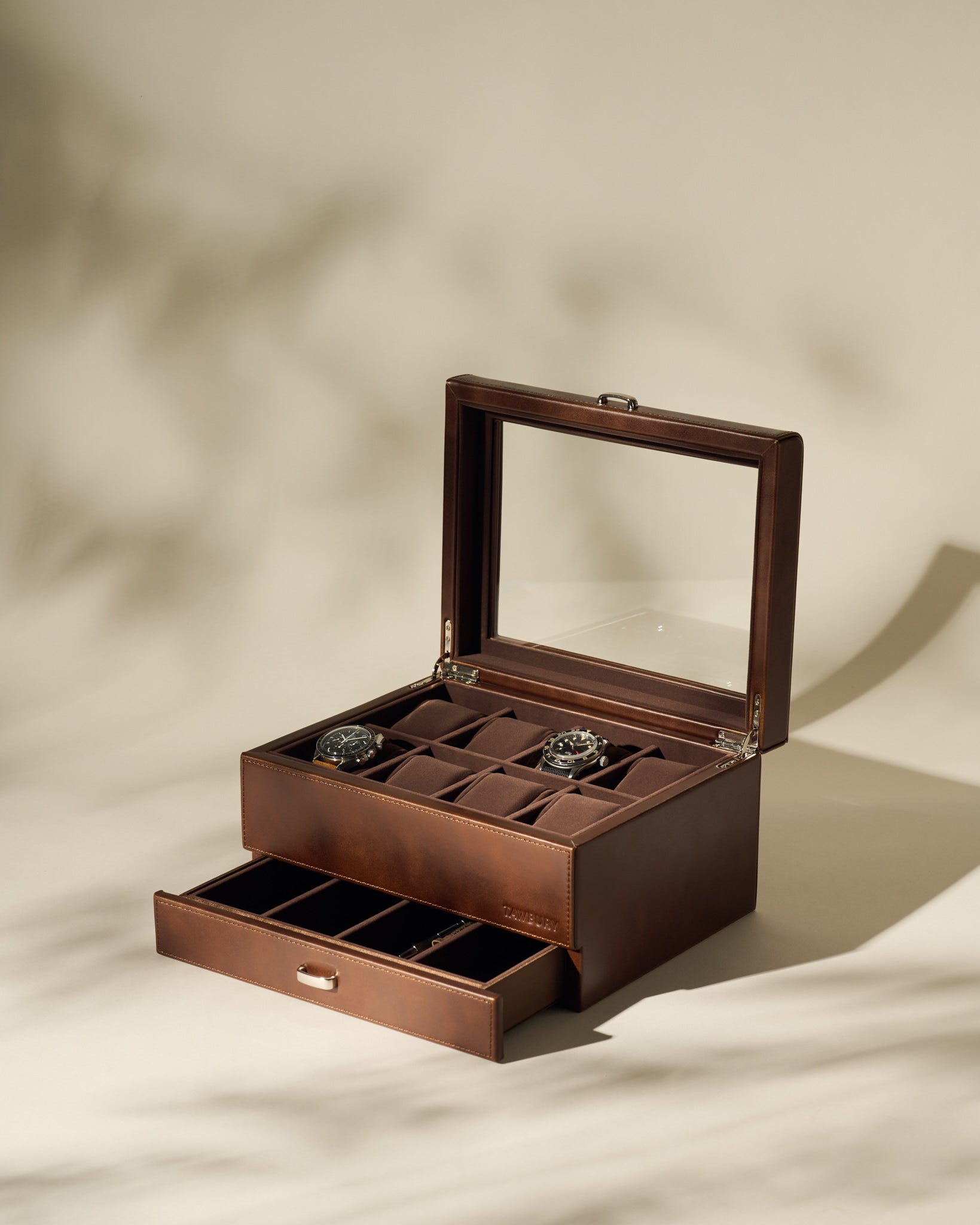 The TAWBURY Bayswater 8 Slot Watch Box with Storage in brown faux leather features a glass lid displaying two watches and a partially open lower drawer, revealing extra compartments for elegant watch organization.