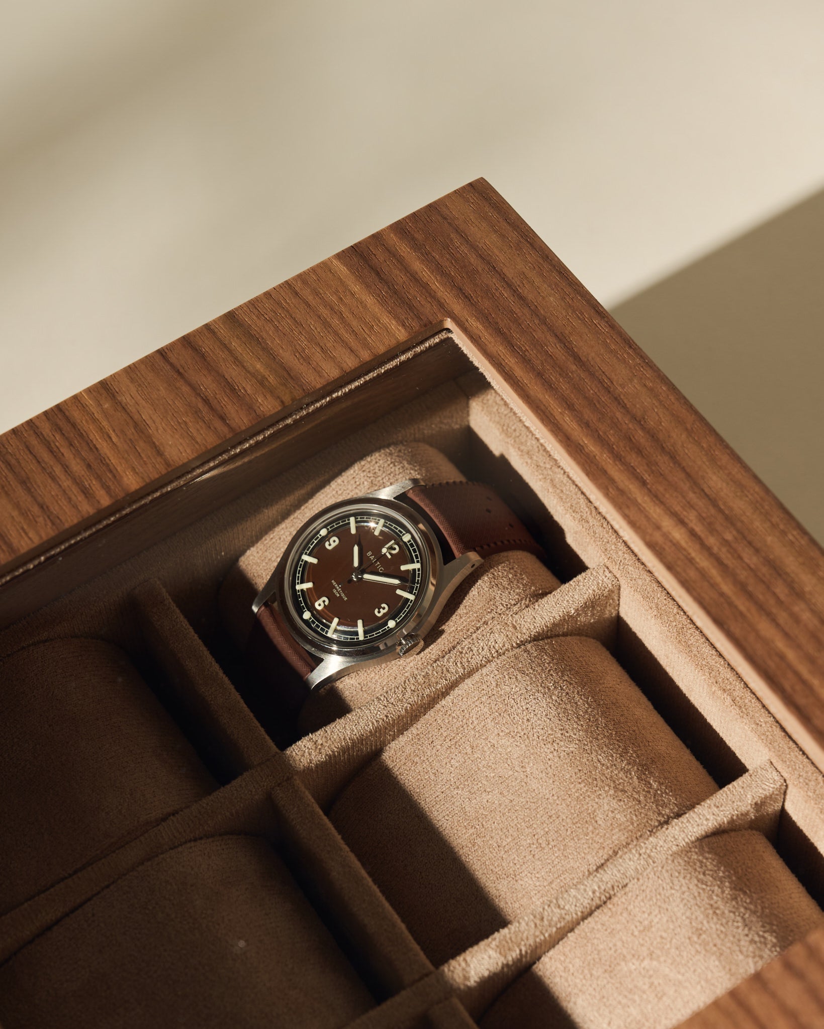 A TAWBURY Grove 6 Slot Wooden Watch Box in light walnut wood with a glass lid stores a wristwatch with brown leather strap and black dial, featuring beige suede compartments to showcase your collection.