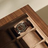 A TAWBURY Grove 6 Slot Wooden Watch Box in light walnut wood with a glass lid stores a wristwatch with brown leather strap and black dial, featuring beige suede compartments to showcase your collection.