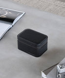 A small black box, specifically the TAWBURY Fraser 2 Watch Travel Case - Black, sitting on a table next to a watch to protect and store watches during travel.