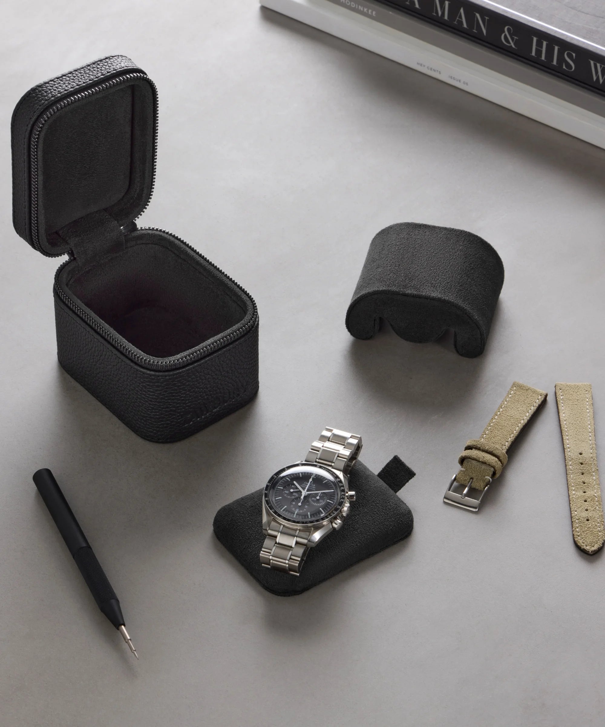 A TAWBURY Fraser 1 Watch Travel Case - Black (Coming Soon) is shown with a wristwatch on a cushion, an extra strap, and a strap-changing tool, all arranged on a grey surface for optimal watch protection.
