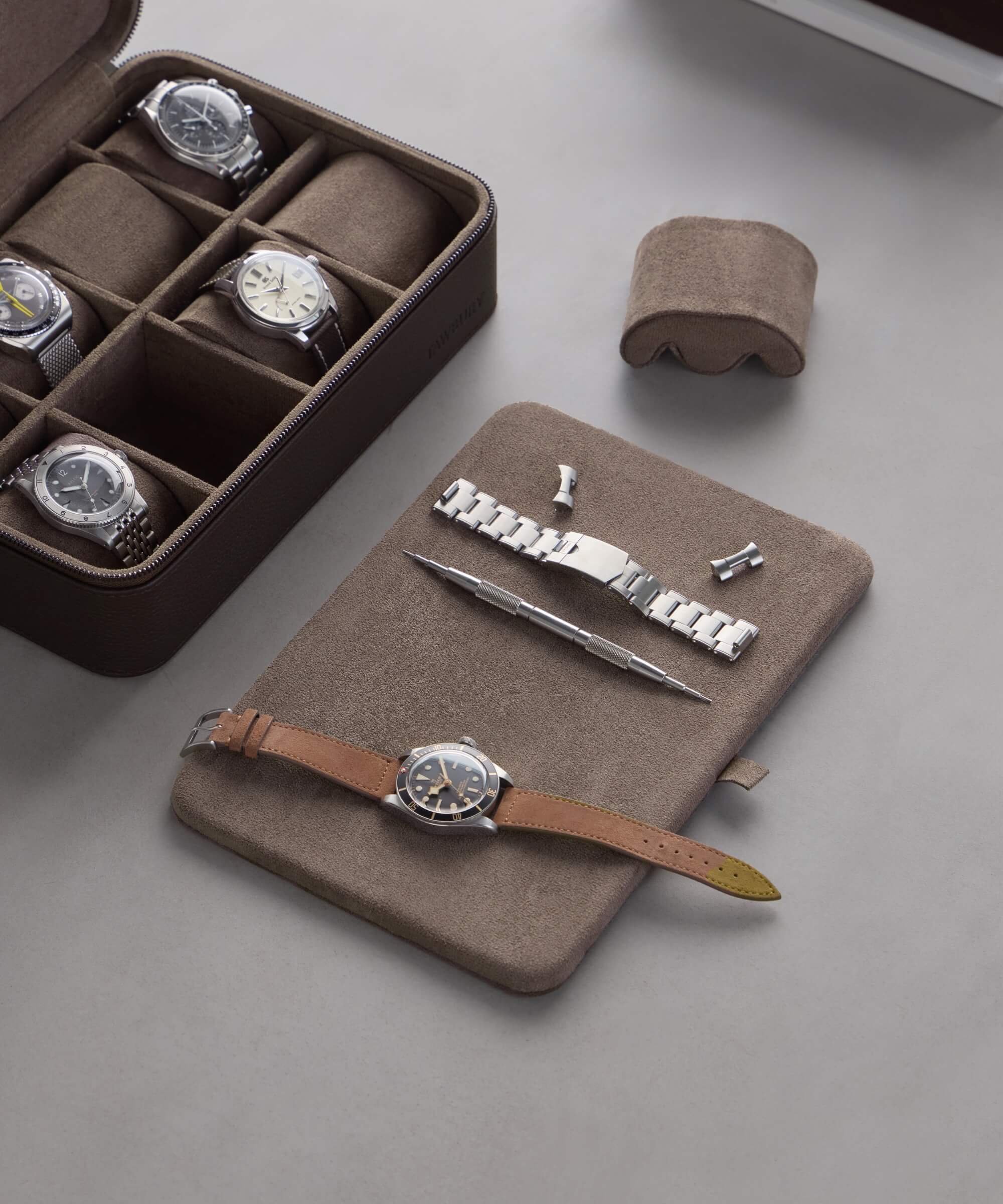 A TAWBURY Fraser 8 Watch Travel Case - Brown is displayed alongside a watch from the Fraser Collection featuring a brown leather strap, paired with a silver bracelet, tool, and watch parts on the gray surface.