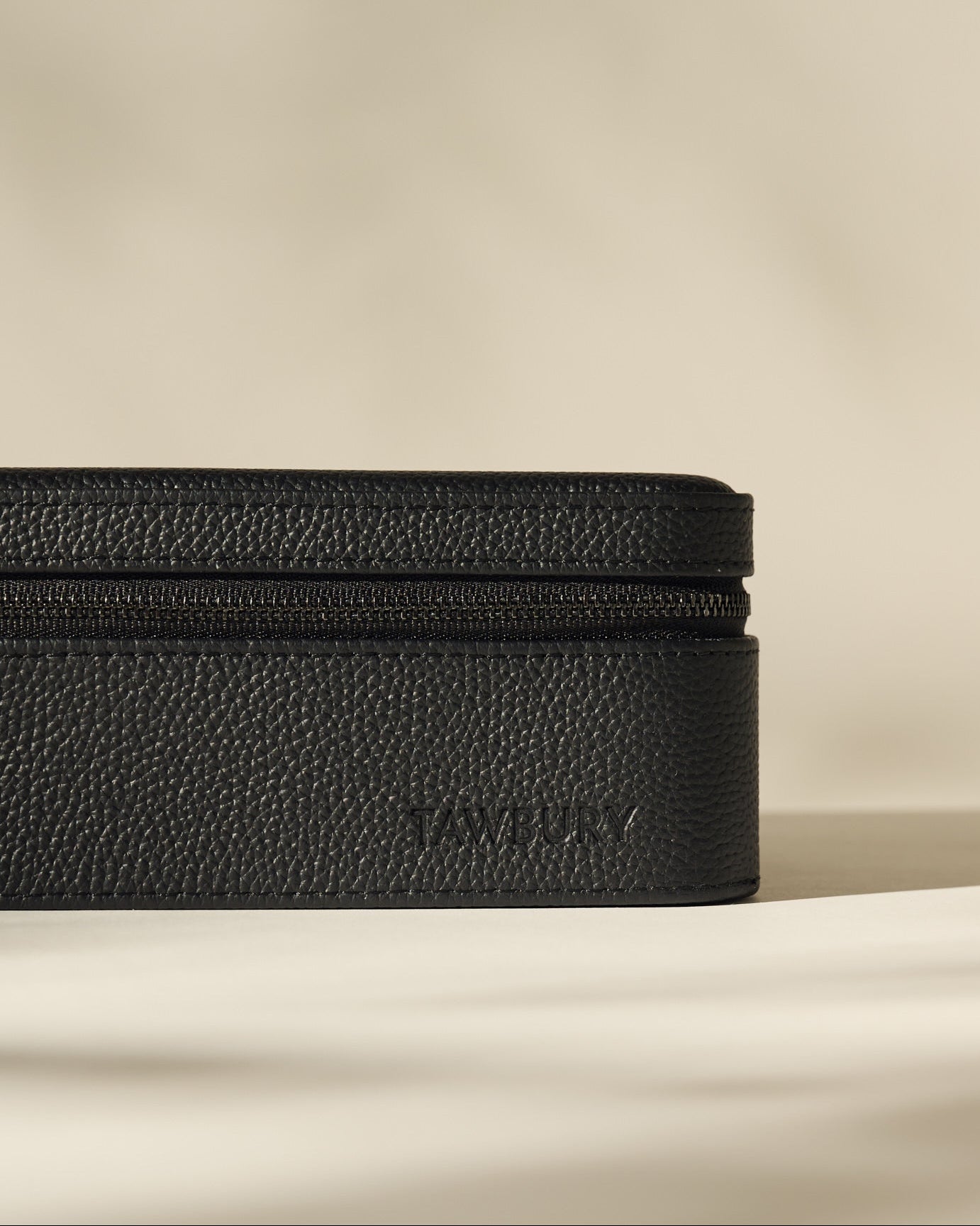 A black textured rectangular leather Fraser 3 Watch Case by TAWBURY, featuring a zippered closure and "TAWBURY" embossed on the side, placed on a plain light surface with a neutral background.