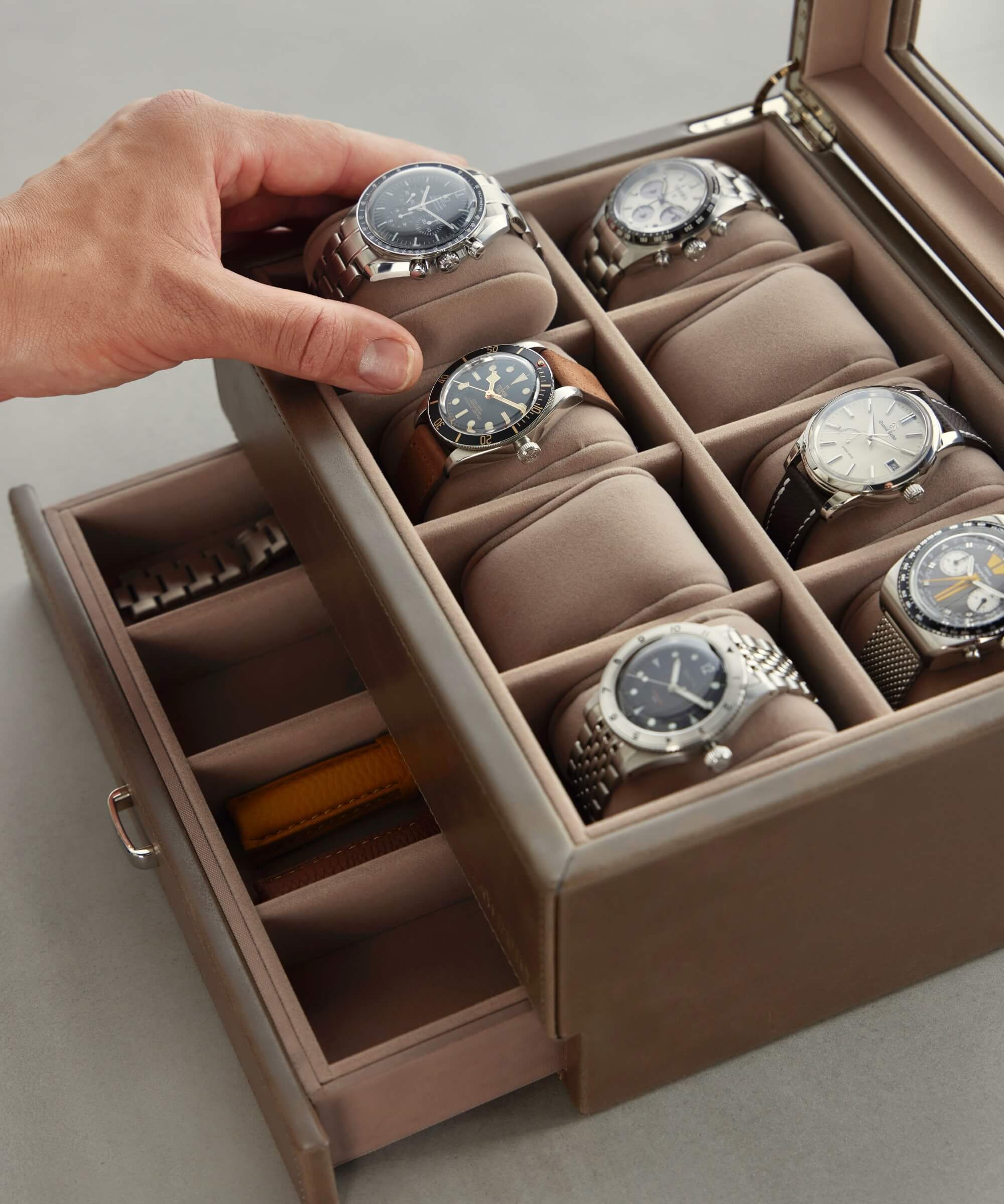 A person's hand is holding a watch in a TAWBURY Bayswater 8 Slot Watch Box with Storage - Taupe, a sophisticated vegan leather case crafted for true watch collectors, elegantly showcasing eight watches with additional storage compartments below.