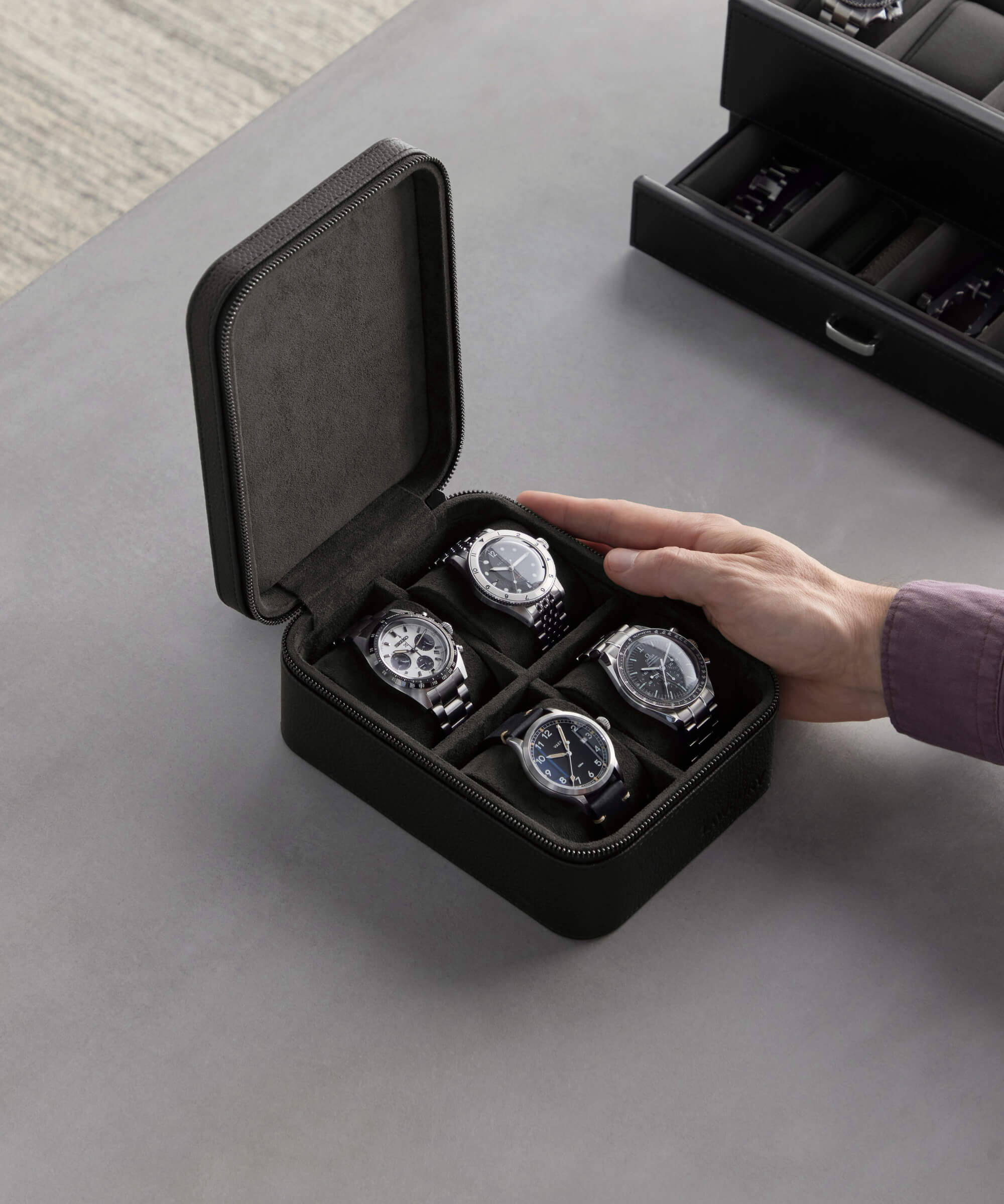 A person in a purple shirt is reaching to open a black leather watch case containing four wristwatches arranged in compartments on a grey surface, showcasing the luxury watch storage of the TAWBURY Fraser 4 Watch Travel Case - Black (Coming Soon).