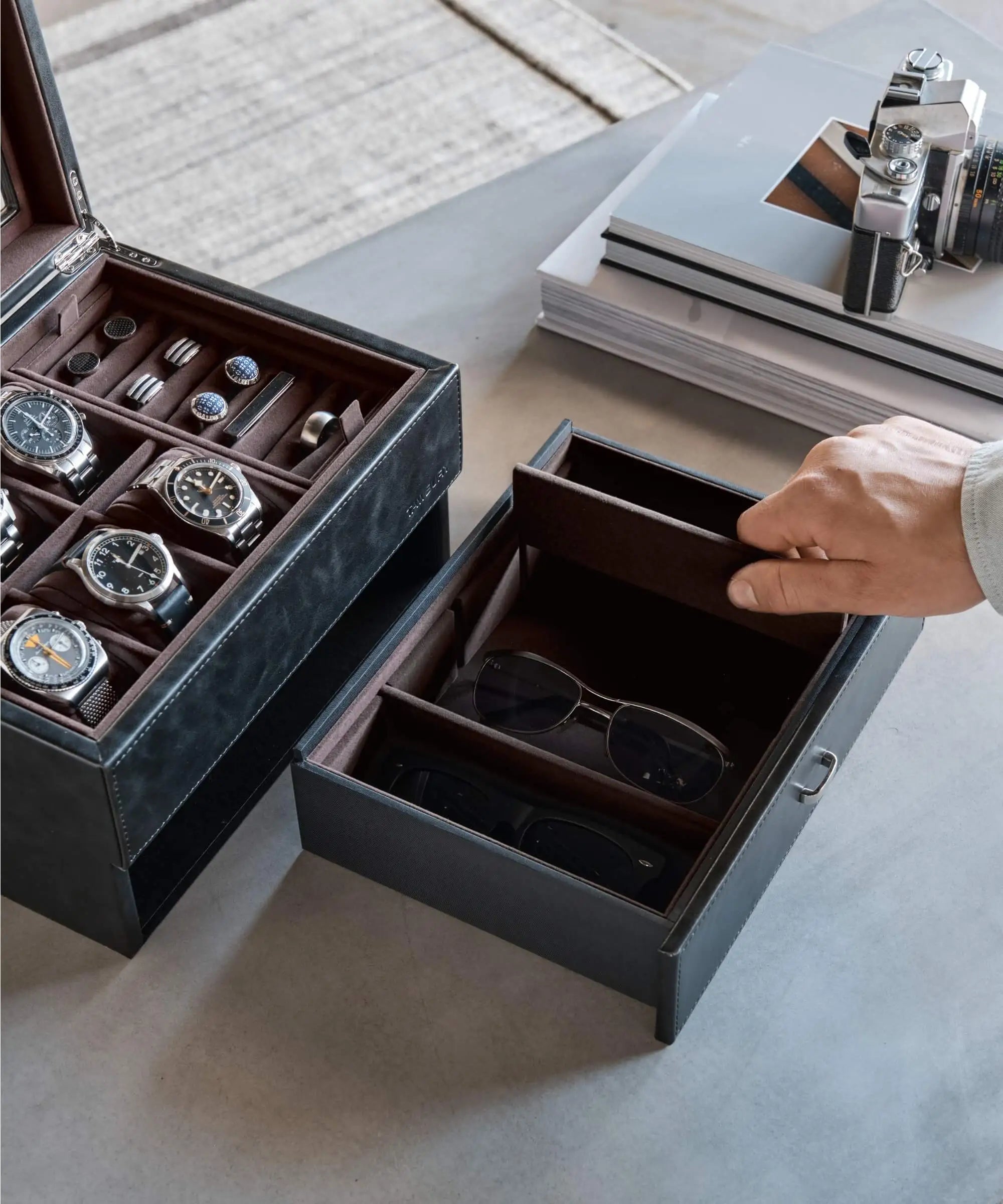 A hand opens a drawer with sunglasses next to the TAWBURY Bayswater 6 Watch Jewellery Box in black/charcoal, filled with watches and cufflinks, with a camera and stacked magazines in the background.