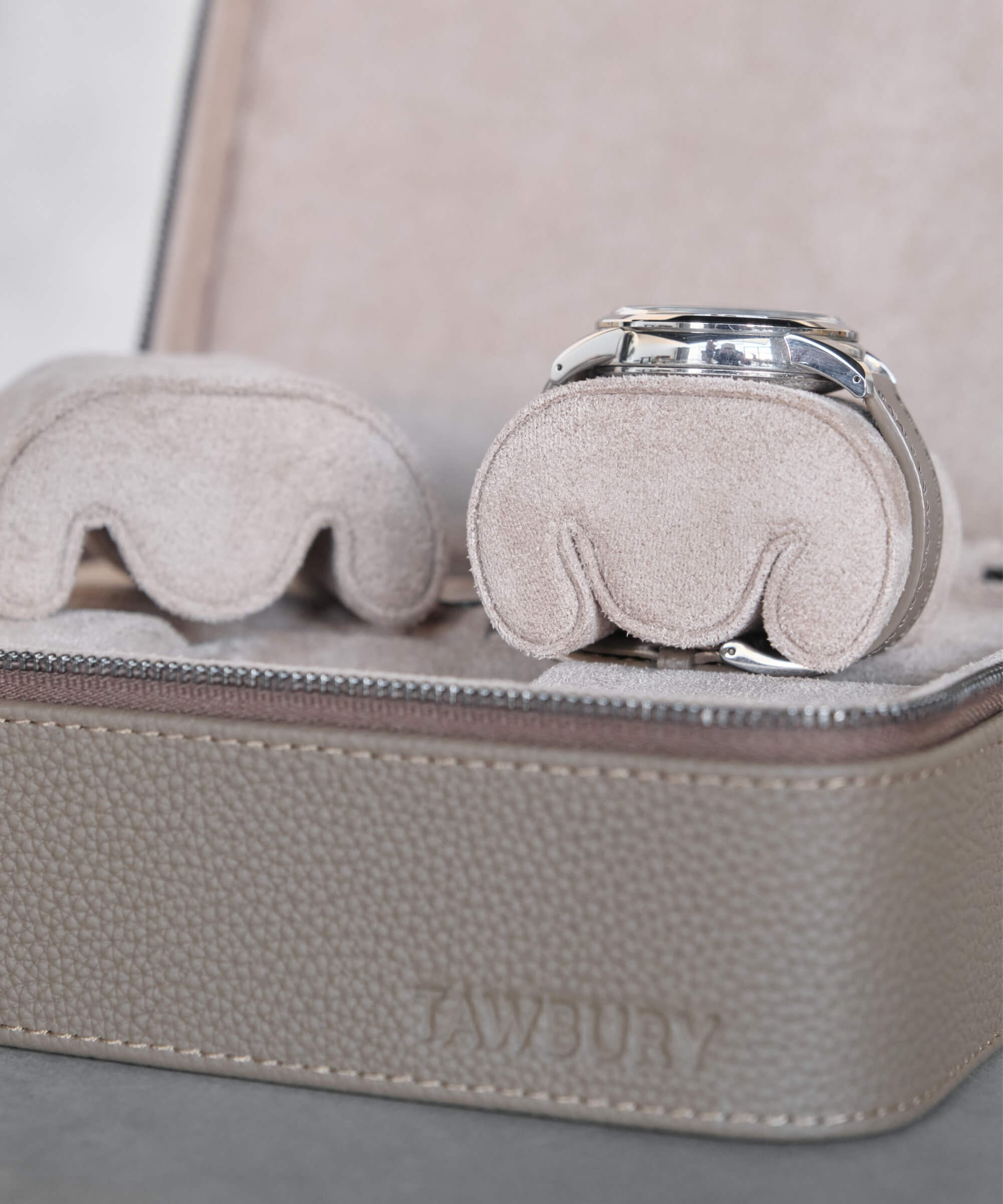 A close-up of a beige watch travel case with the brand name "TAWBURY" embossed on it. Inside, the wristwatch is secured on one of the slender Fraser Replacement Watch Case Pillows - X-Small - Taupe/Cream, ensuring maximum protection for your timepiece.