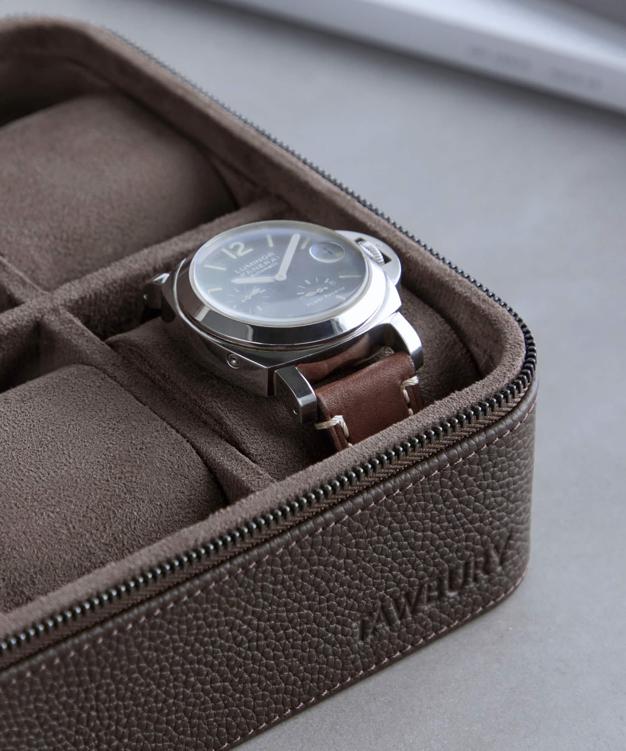 A wristwatch with a brown leather strap rests inside a TAWBURY Fraser 4 Watch Travel Case - Brown (Coming Soon) with a zipper. The elegant case, ideal for watch storage, is thoughtfully divided into compartments.