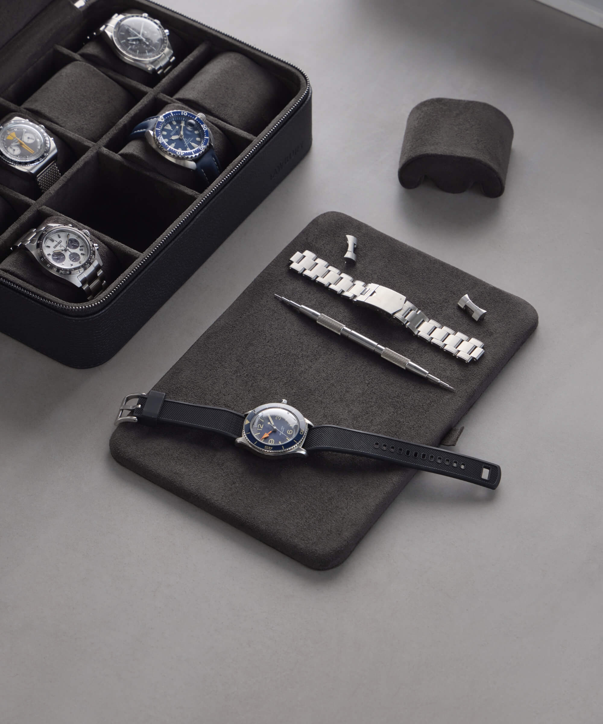 Watches and accessories are elegantly displayed with a TAWBURY Fraser 8 Watch Travel Case in black. A black watch sits on a pad with replacement parts, complemented by tools and extra bands, completing this refined setup.