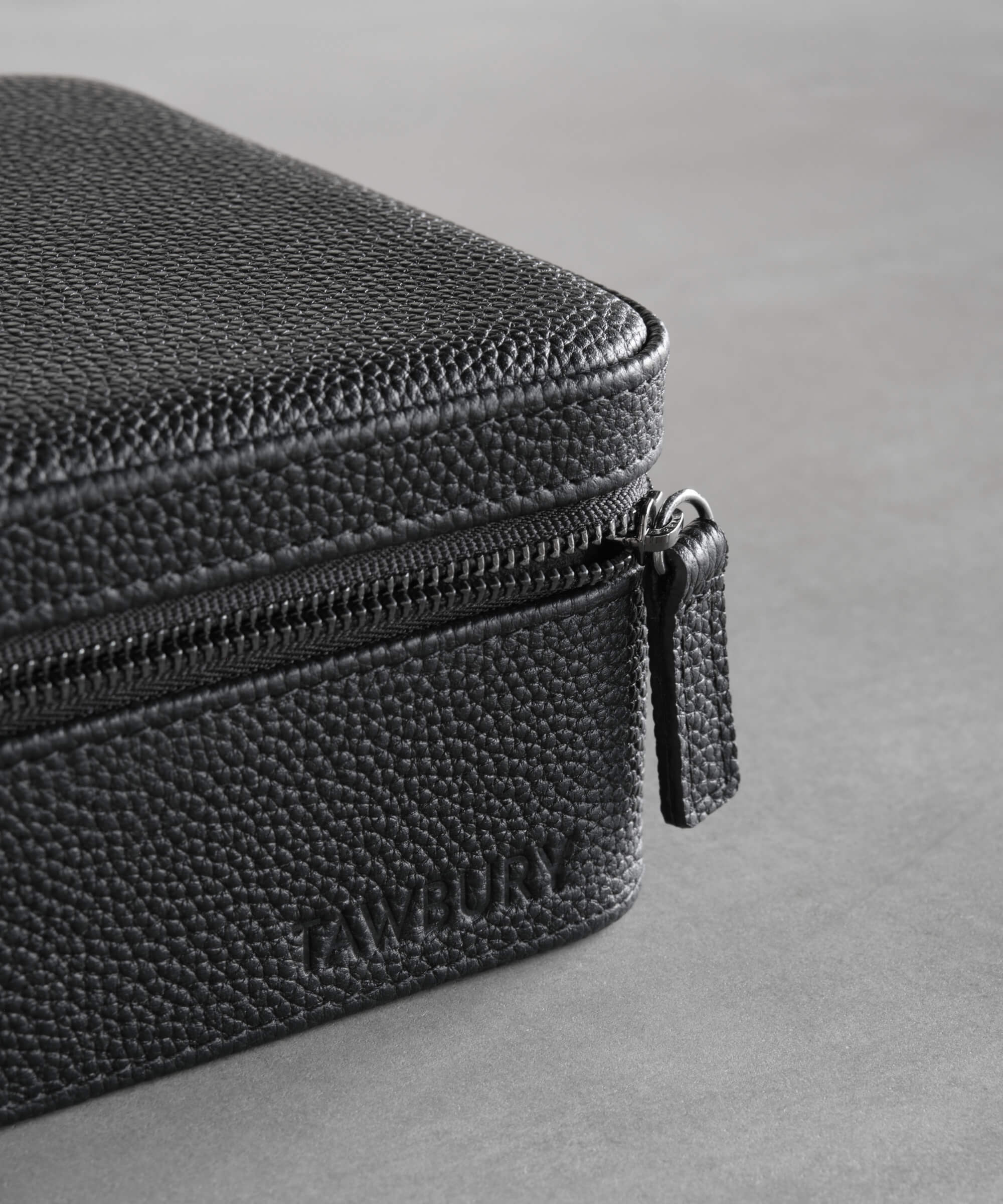 Close-up of a black textured leather case with a visible zipper and the embossed brand name "Tawbury" on the front, part of the luxury TAWBURY range. This Fraser 6 Watch Travel Case - Black (Coming Soon) epitomizes elegance and craftsmanship.