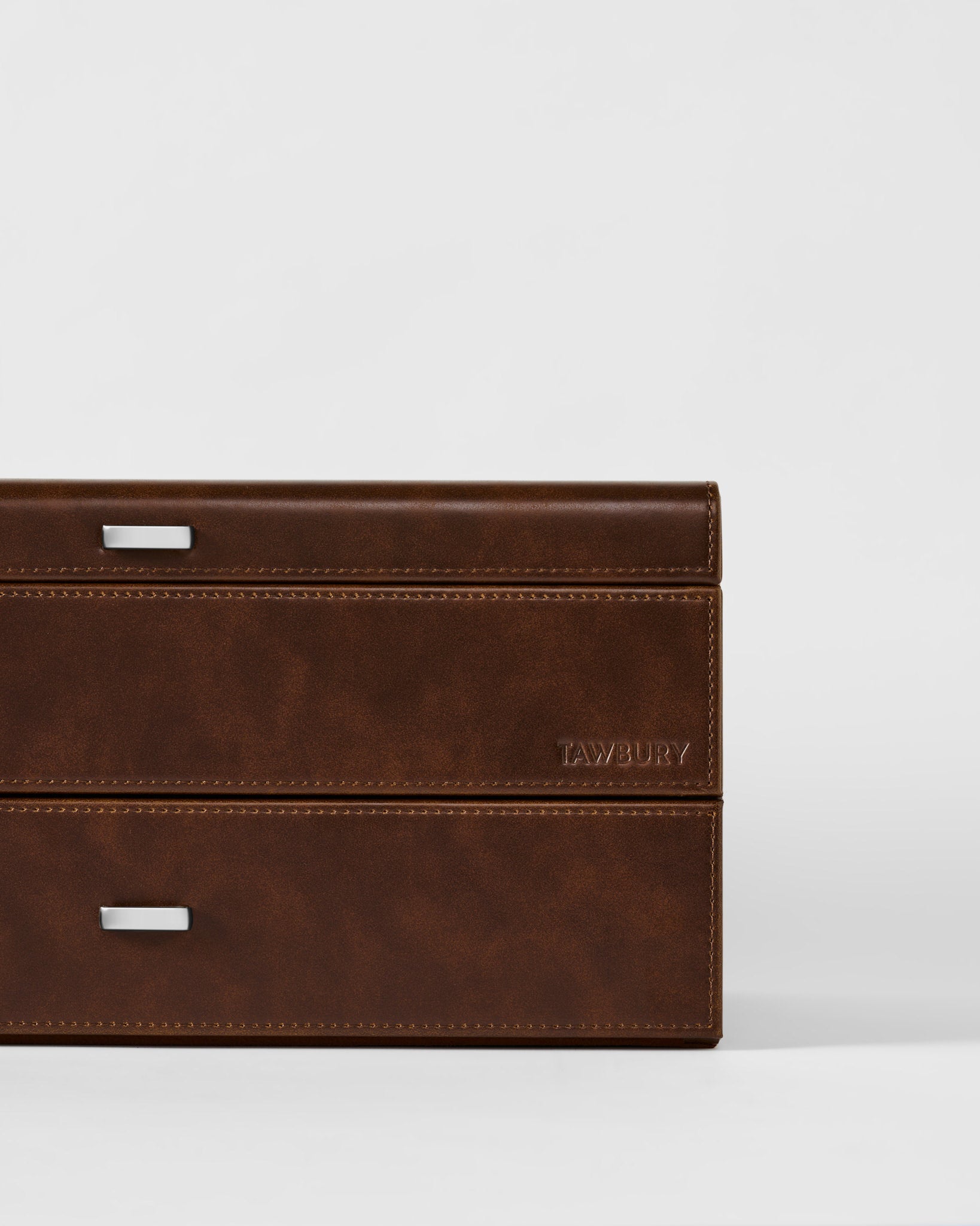A close-up of the TAWBURY Bayswater 24 Slot Watch Box with Drawer in brown leather, featuring metal handles and “TAWBURY” embossed on the side, set against a plain white background.