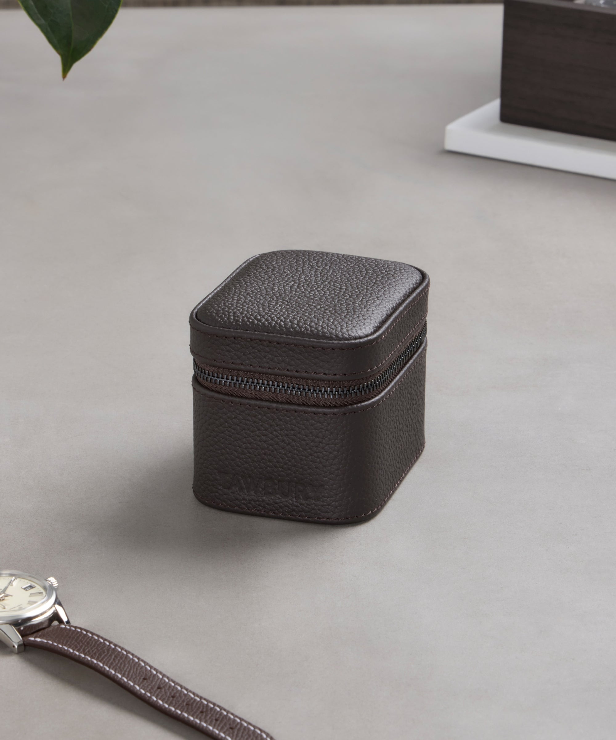 The TAWBURY Fraser 1 Watch Travel Case - Brown (Coming Soon) rests on a grey surface beside a wristwatch and white tray, providing stylish protection for your timepiece.