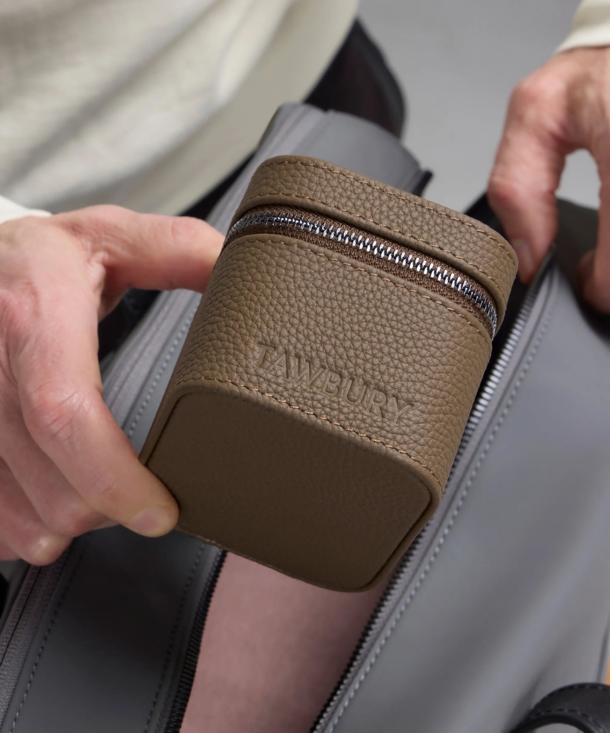 Someone holds the TAWBURY Fraser 1 Watch Travel Case - Taupe (Coming Soon), a compact, textured brown zippered case with "TAWBURY" embossed on it, above an open gray bag.