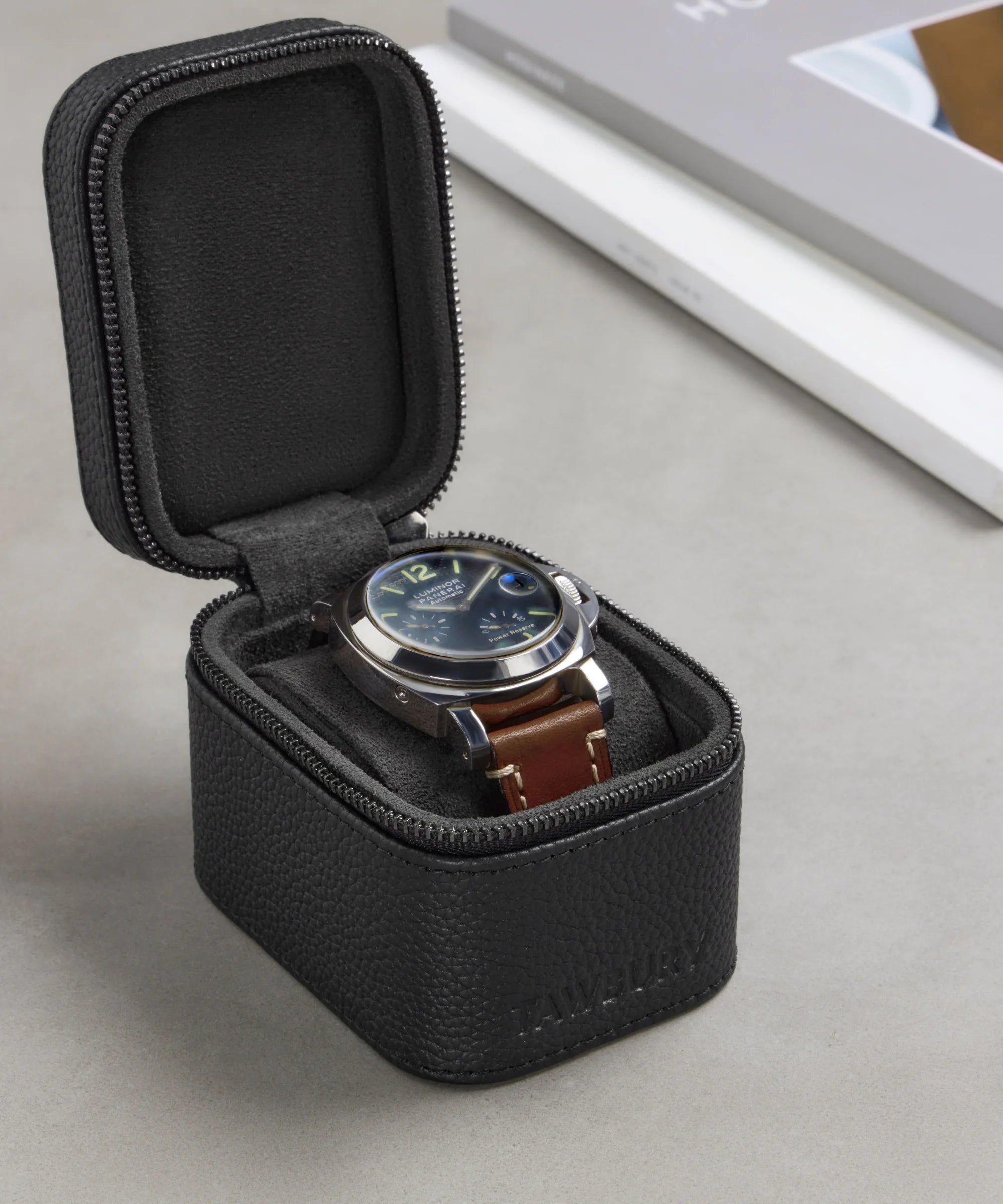 A wristwatch with a brown leather strap is displayed in the open Fraser 1 Watch Travel Case - Black by TAWBURY on a gray surface.