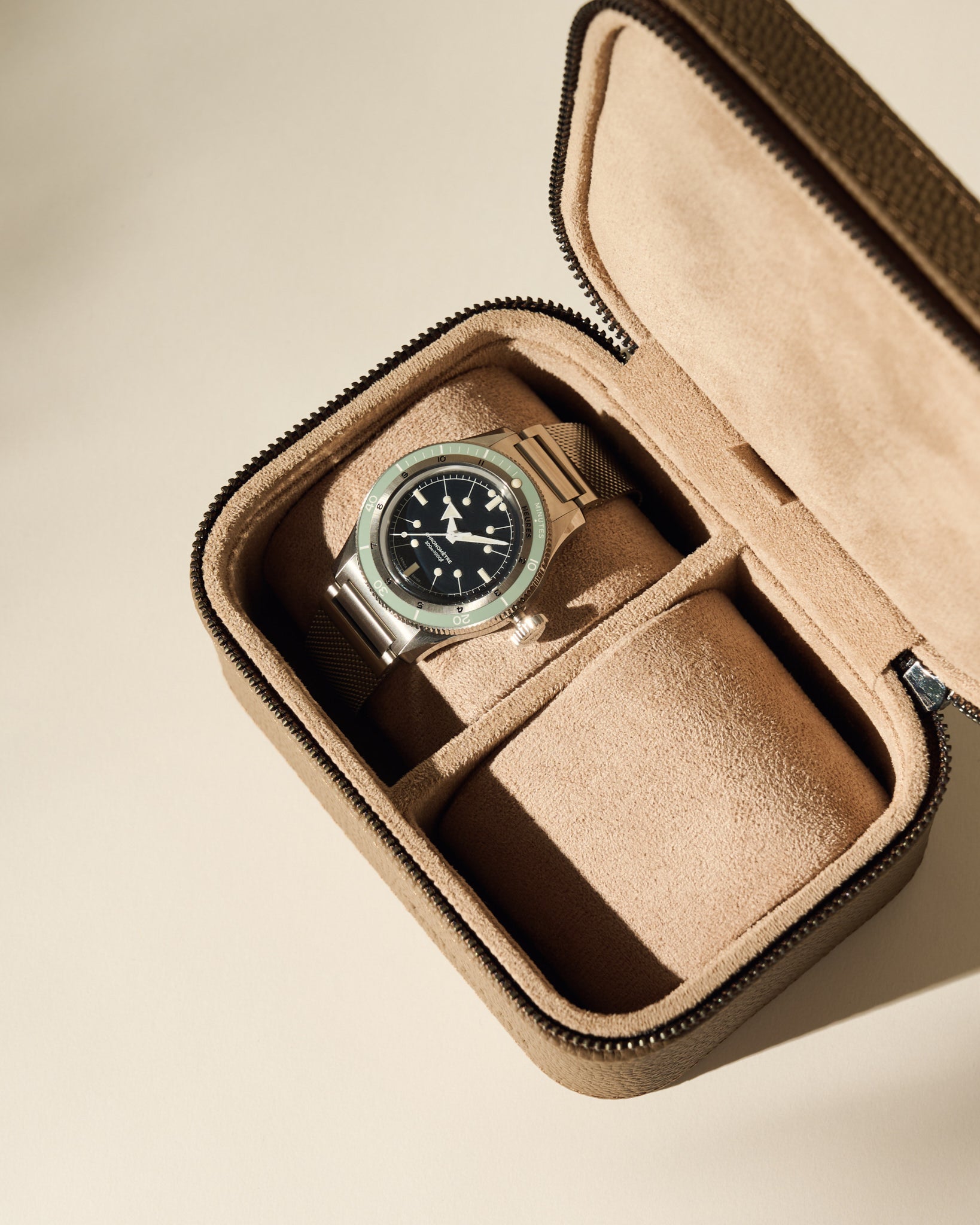 A wristwatch with a black face and metal strap is displayed inside the open TAWBURY Fraser 2 Watch Case in taupe.