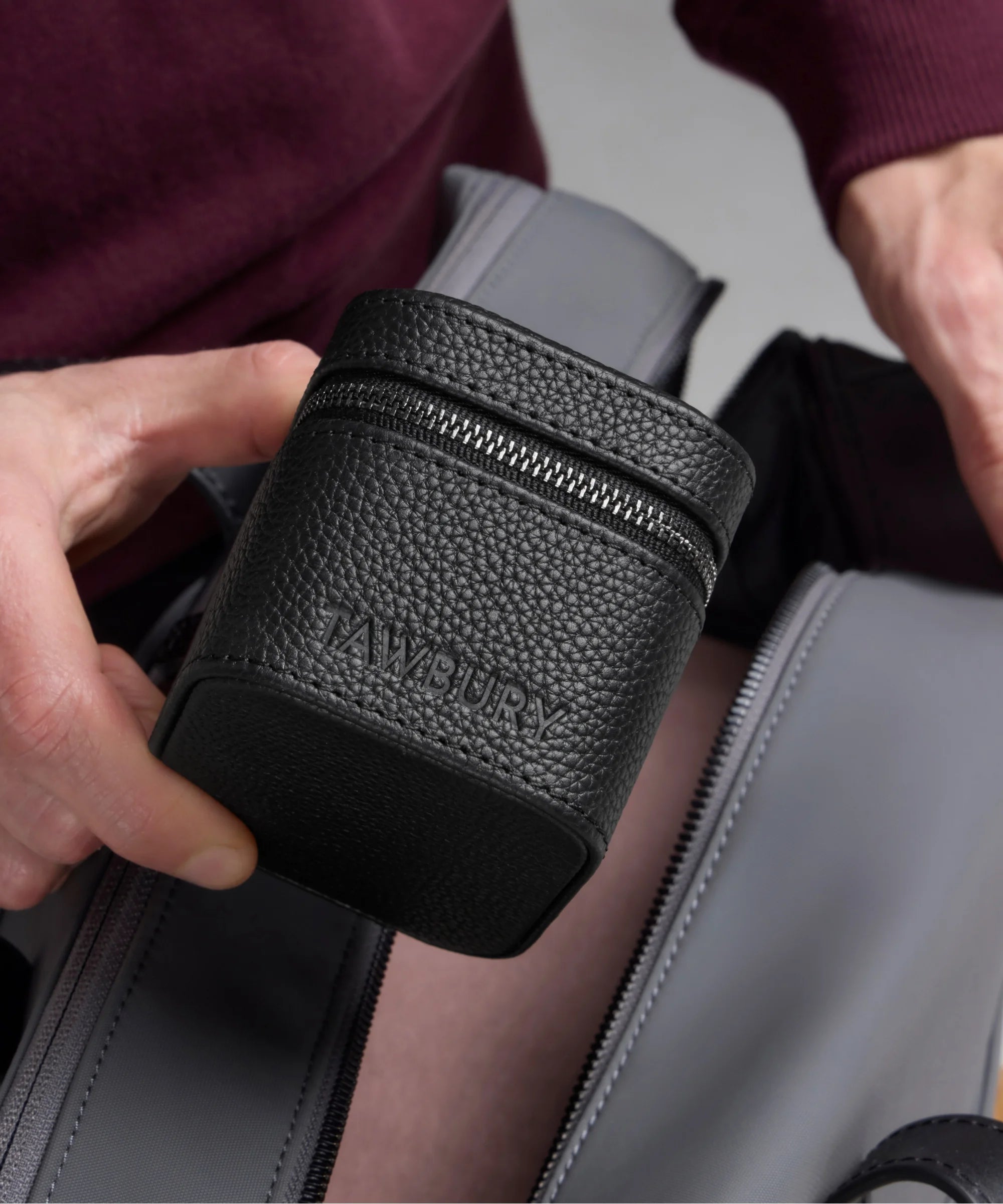 A person holds the Fraser 1 Watch Travel Case - Black by TAWBURY, featuring a textured leather zippered design, above an open gray bag.