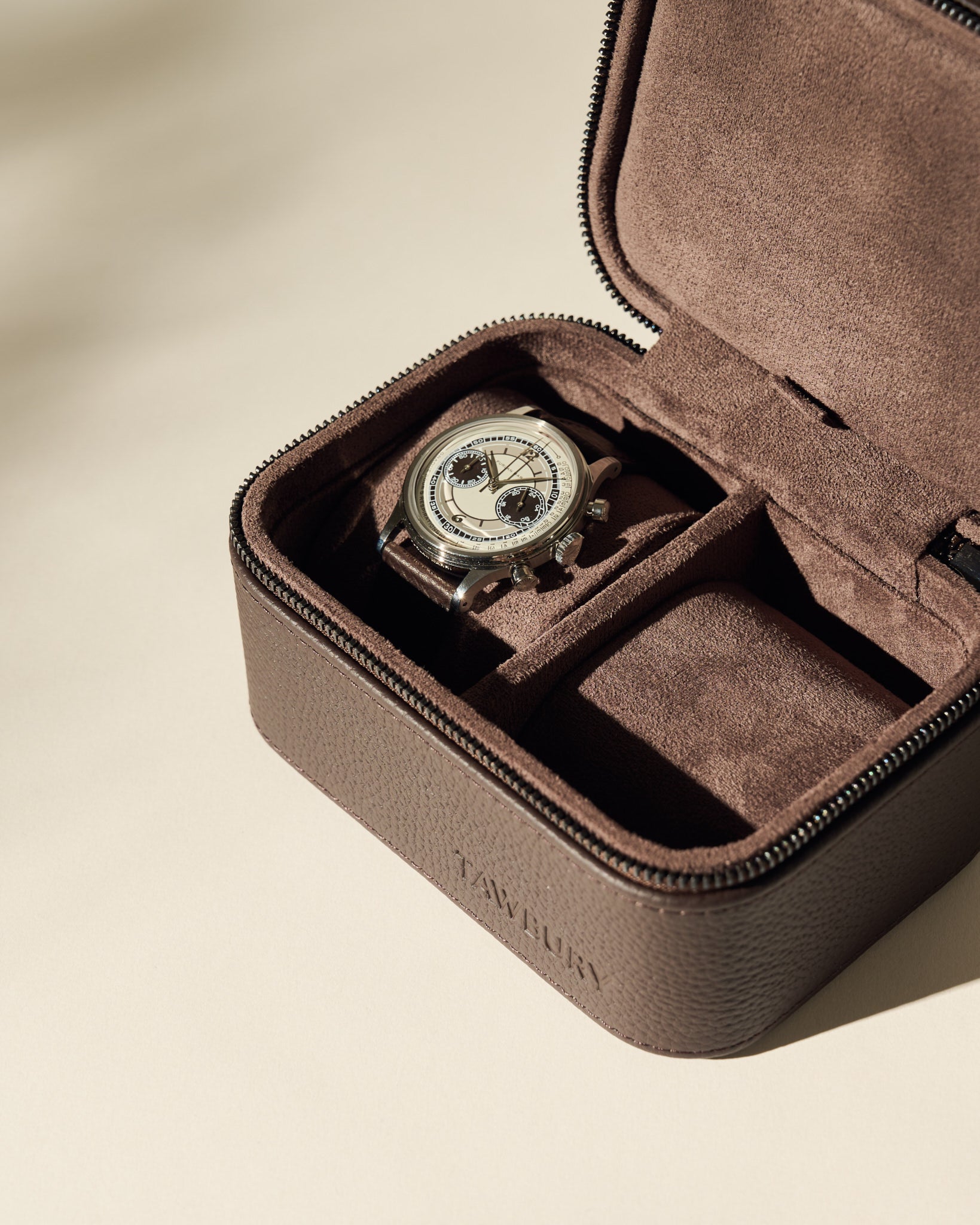 A wristwatch with a light dial and dark strap is displayed inside an open TAWBURY Fraser 2 Watch Case in brown, featuring a suede interior.