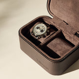A wristwatch with a light dial and dark strap is displayed inside an open TAWBURY Fraser 2 Watch Case in brown, featuring a suede interior.