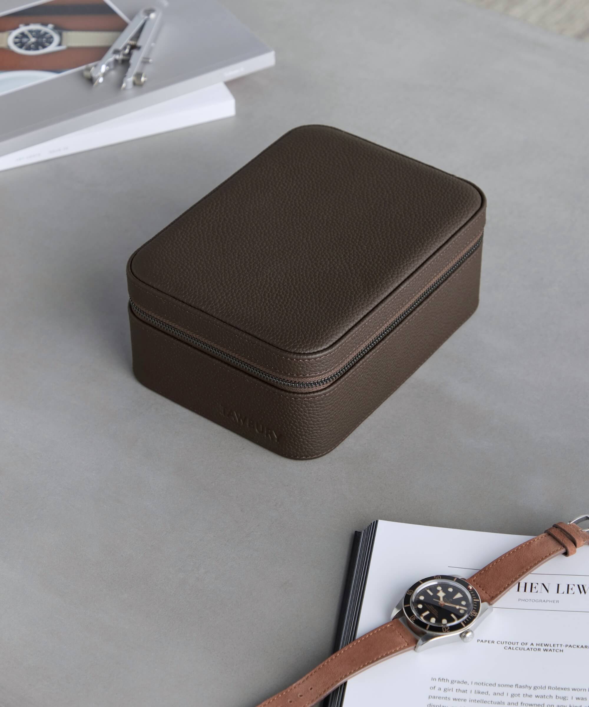 A brown Fraser 4 Watch Travel Case - Brown (Coming Soon) rests on a light gray surface with an open book nearby. A wristwatch and a pair of eyeglasses lie in the background, showcasing the elegance of the TAWBURY range.