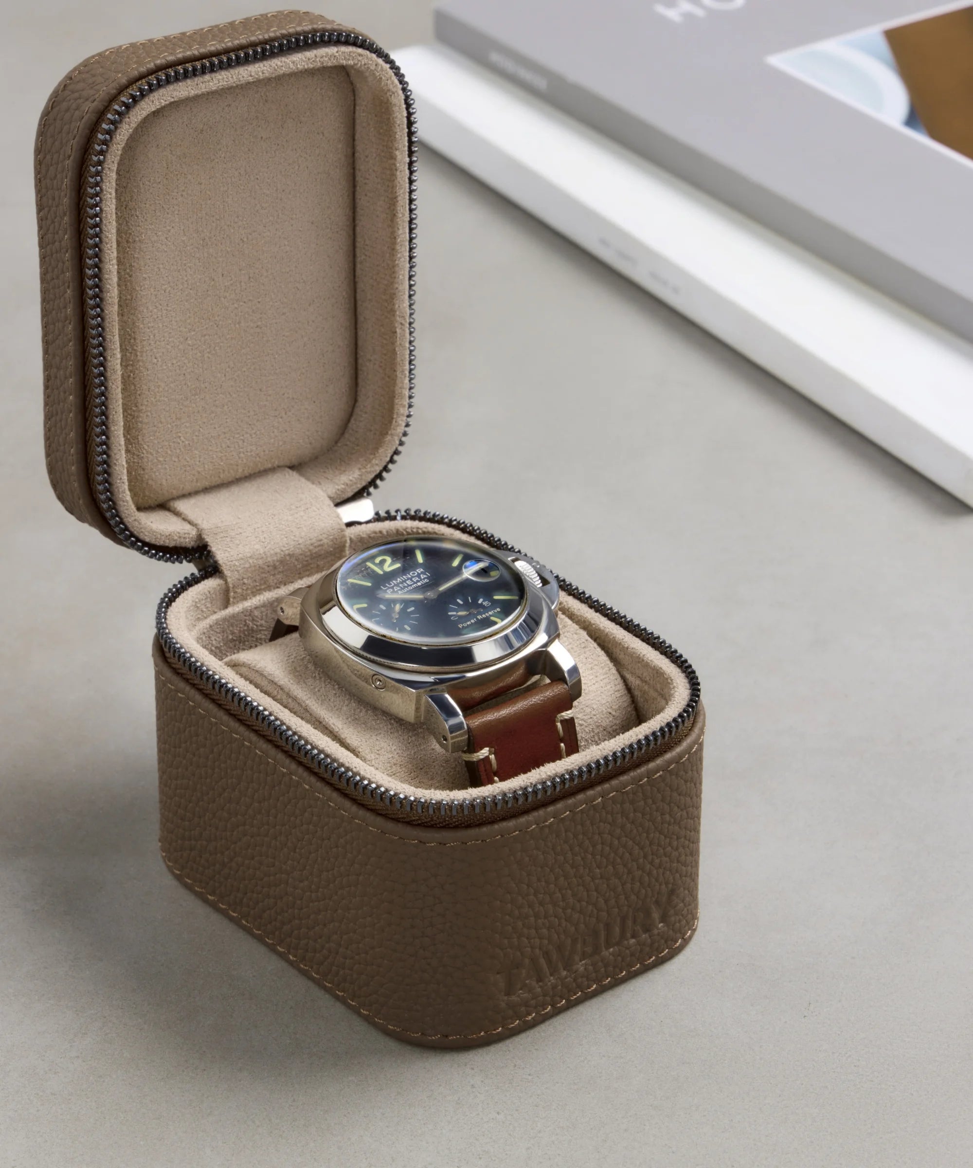 A wristwatch with a brown leather strap is displayed in an open TAWBURY Fraser 1 Watch Travel Case - Taupe (Coming Soon) on a gray surface, with part of a book visible in the background.