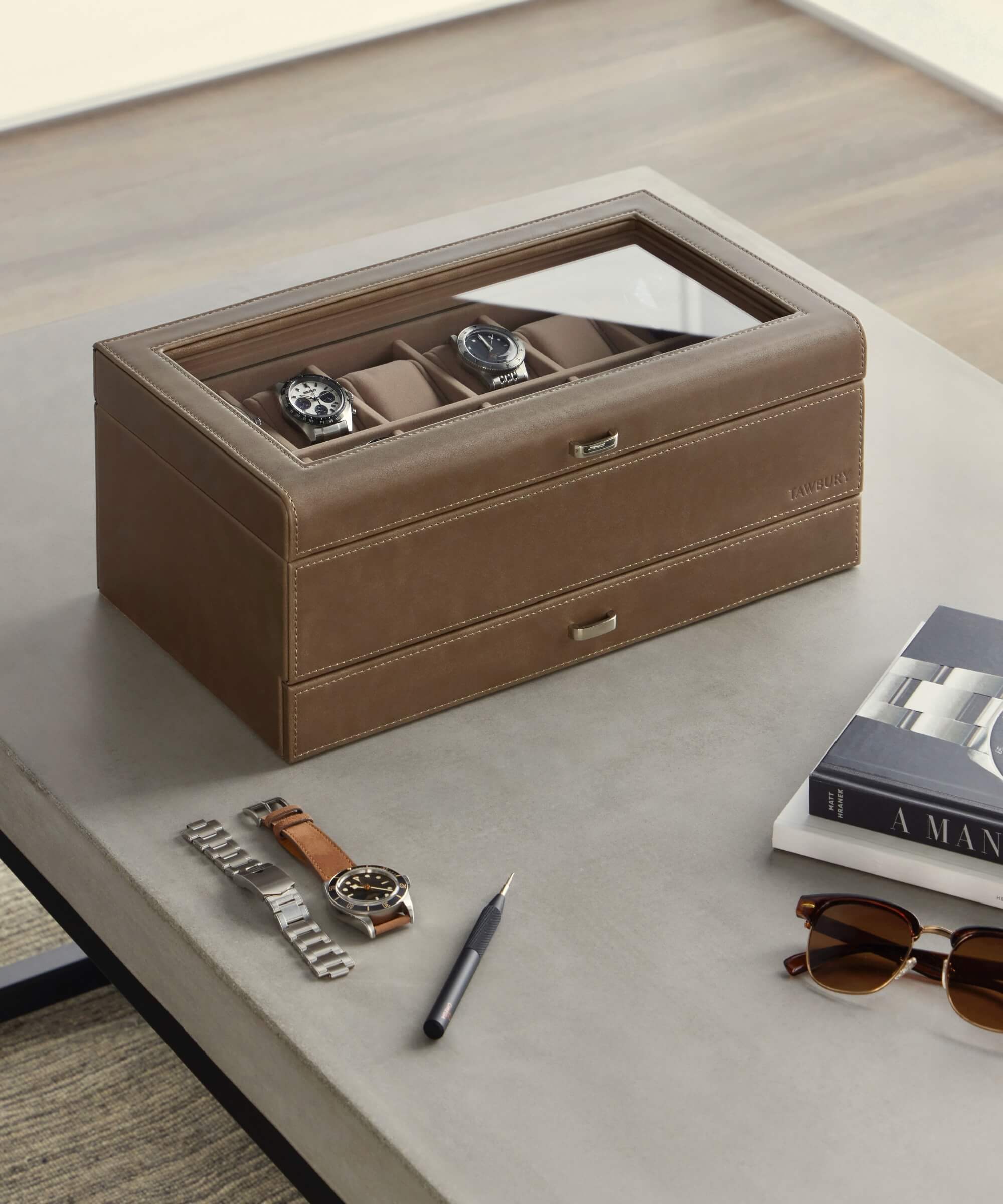 On the table sits a stylish TAWBURY Bayswater 12 Slot Watch Box with Storage in taupe vegan leather, featuring a glass top that beautifully displays three exquisite watches. Nearby, a pen, sunglasses, books, and a metal watch bracelet complete this elegant arrangement of watch collector accessories.