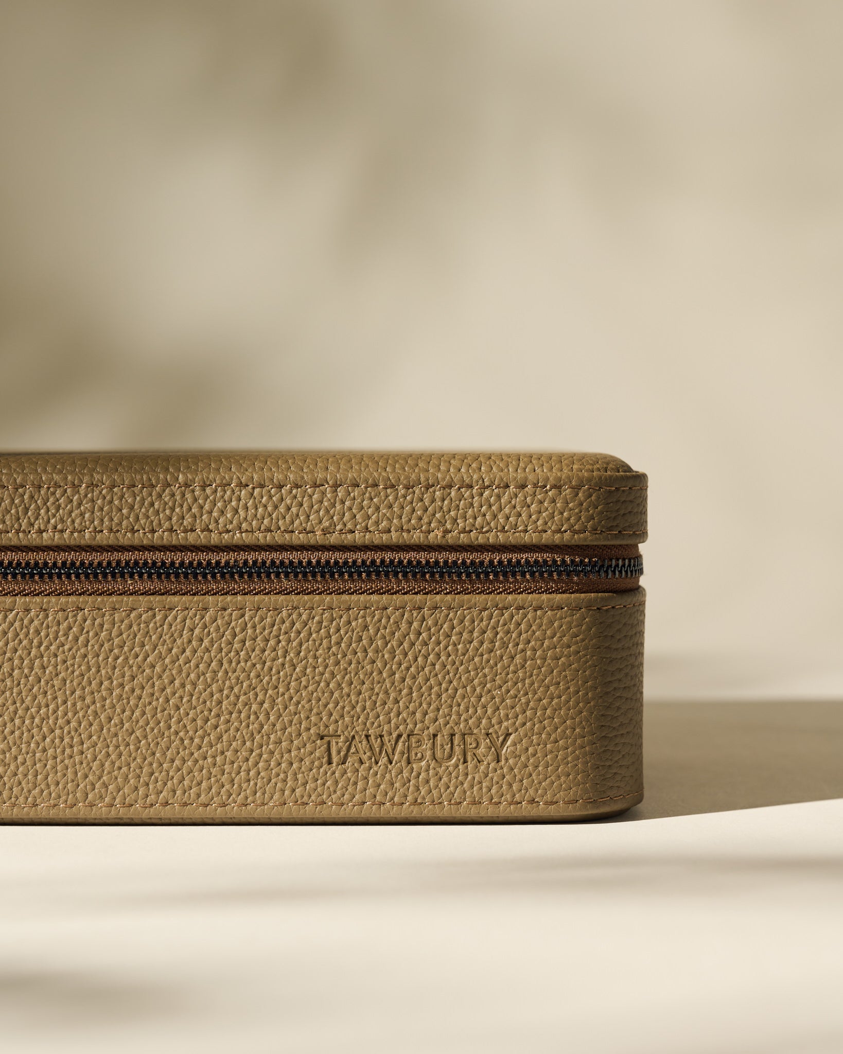 The Fraser 3 Watch Case with Storage by TAWBURY, in taupe textured leather and embossed with the brand name on the front, rests on a flat surface against a neutral background.