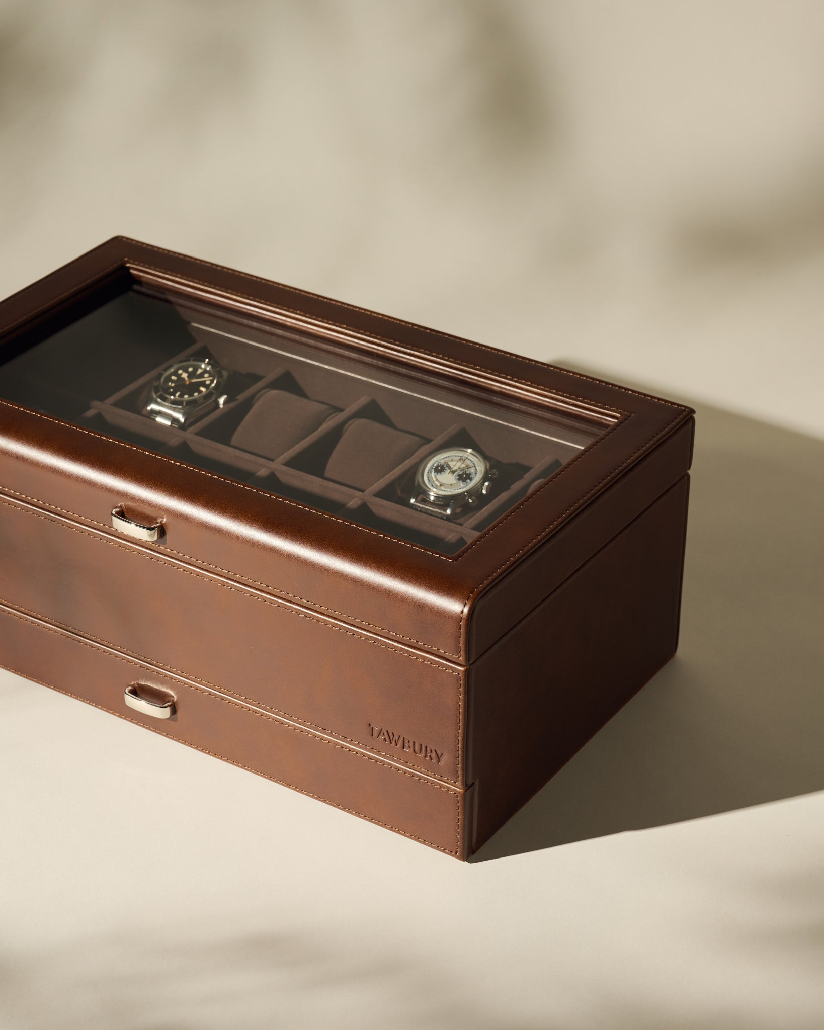 The TAWBURY Bayswater 12 Slot Watch Box with Storage in brown leather features a glass top and showcases two wristwatches inside, set on a neutral surface with soft shadows.