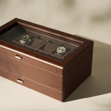 The TAWBURY Bayswater 12 Slot Watch Box with Storage in brown leather features a glass top and showcases two wristwatches inside, set on a neutral surface with soft shadows.