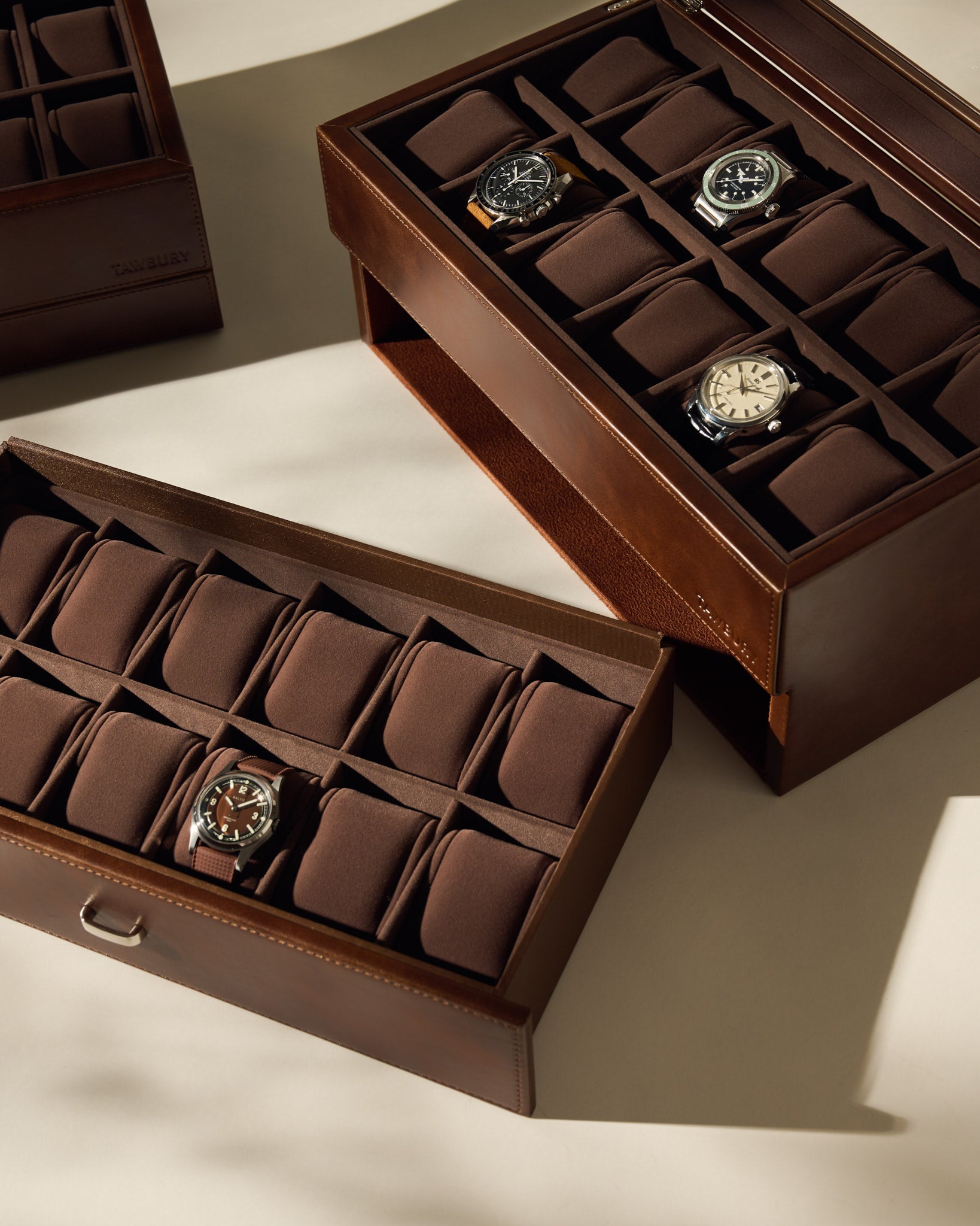 The TAWBURY Bayswater 24 Slot Watch Box with Drawer in brown offers elegant storage for multiple watches, displayed on a beige surface under soft lighting.