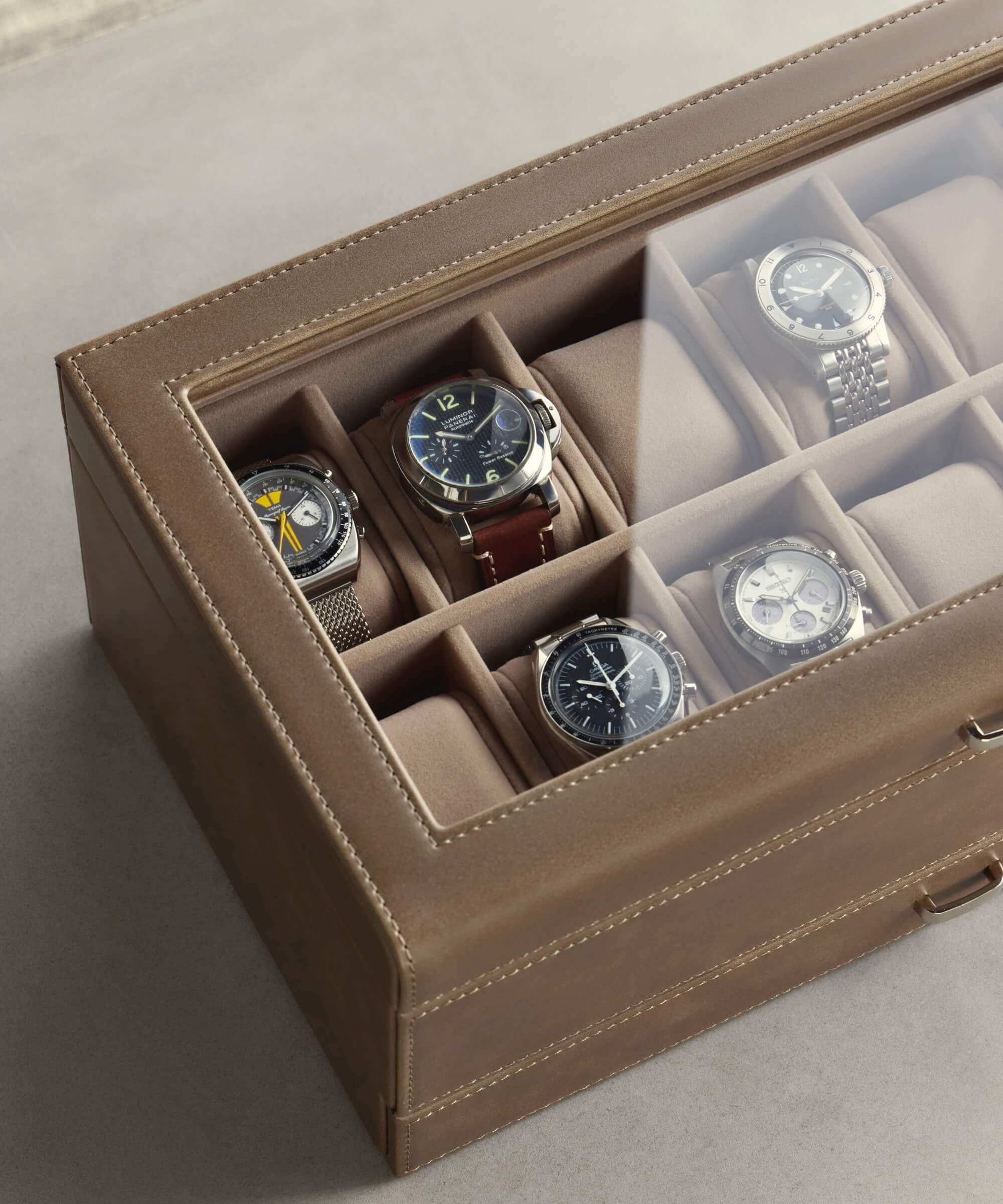 The TAWBURY Bayswater 12 Slot Watch Box with Storage in Taupe is a chic option made from vegan leather. It features a transparent lid that elegantly showcases twelve wristwatches, each securely nestled in its own padded compartment—ideal for watch enthusiasts.