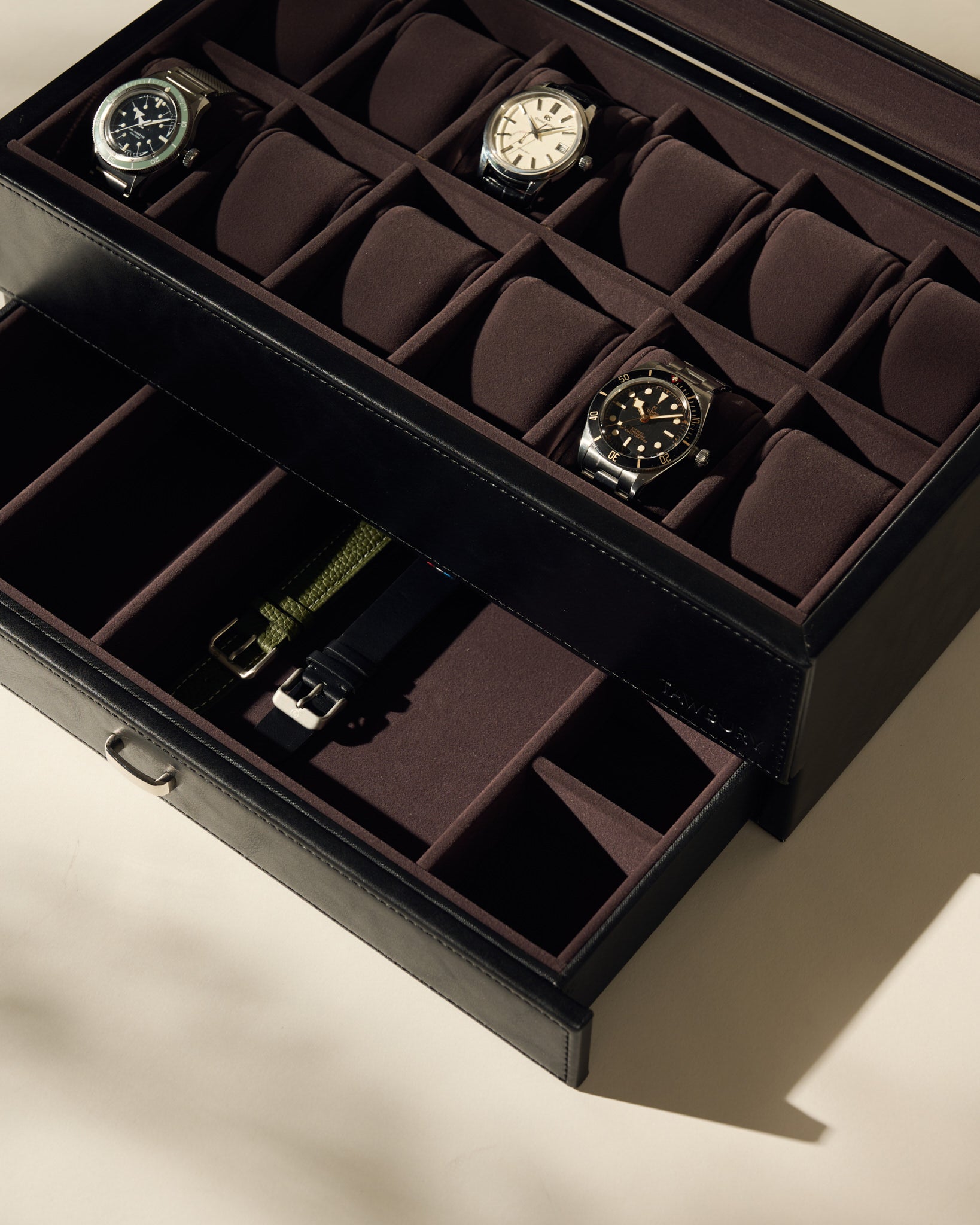 The TAWBURY Bayswater 12 Slot Watch Box with Storage in Black/Charcoal displays three watches in the top row, with empty compartments below, all photographed under soft lighting.