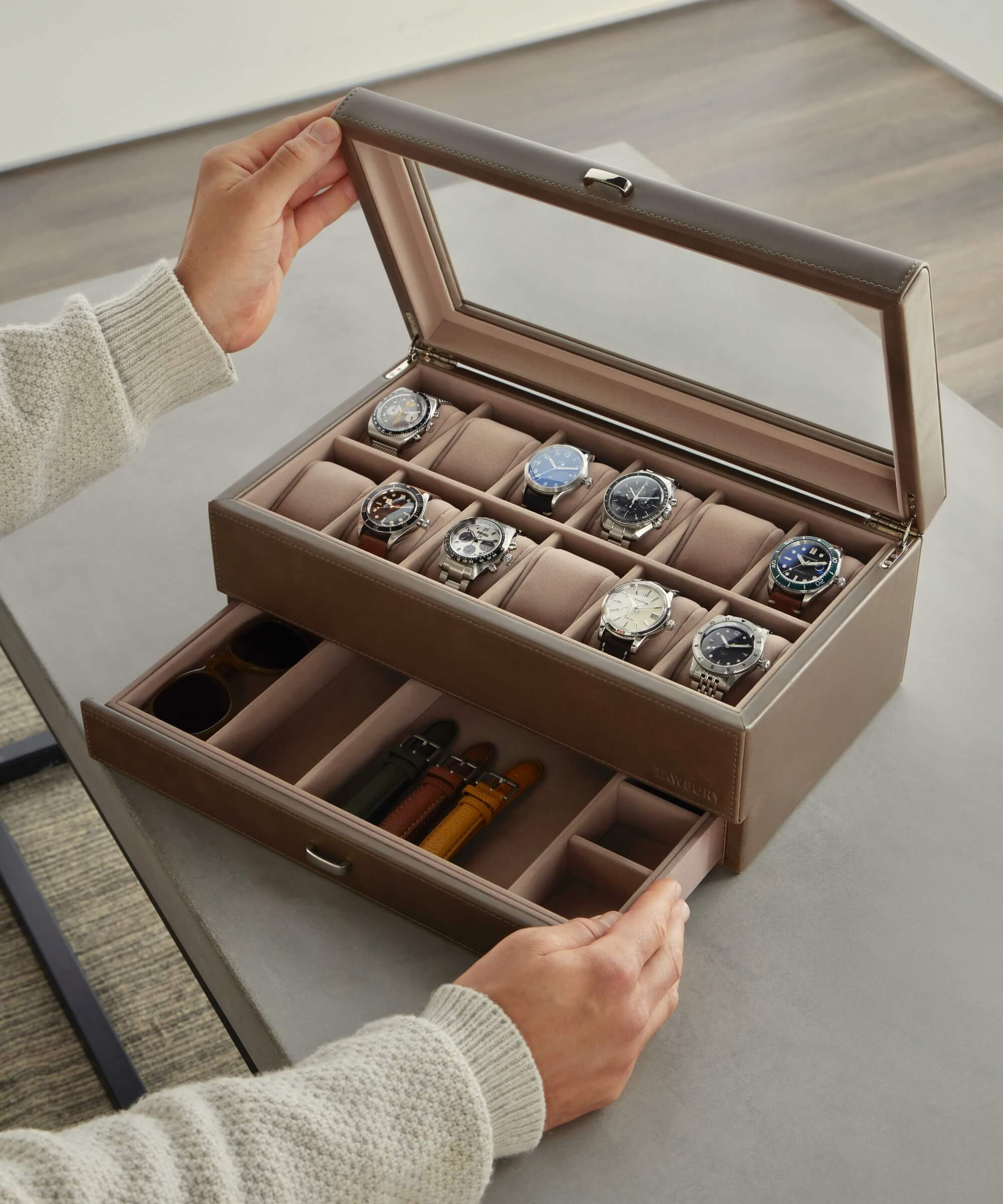 A person opens the TAWBURY Bayswater 12 Slot Watch Box with Storage in taupe, revealing nine watches resting on plush watch pillows and three watch straps organized in dedicated compartments.