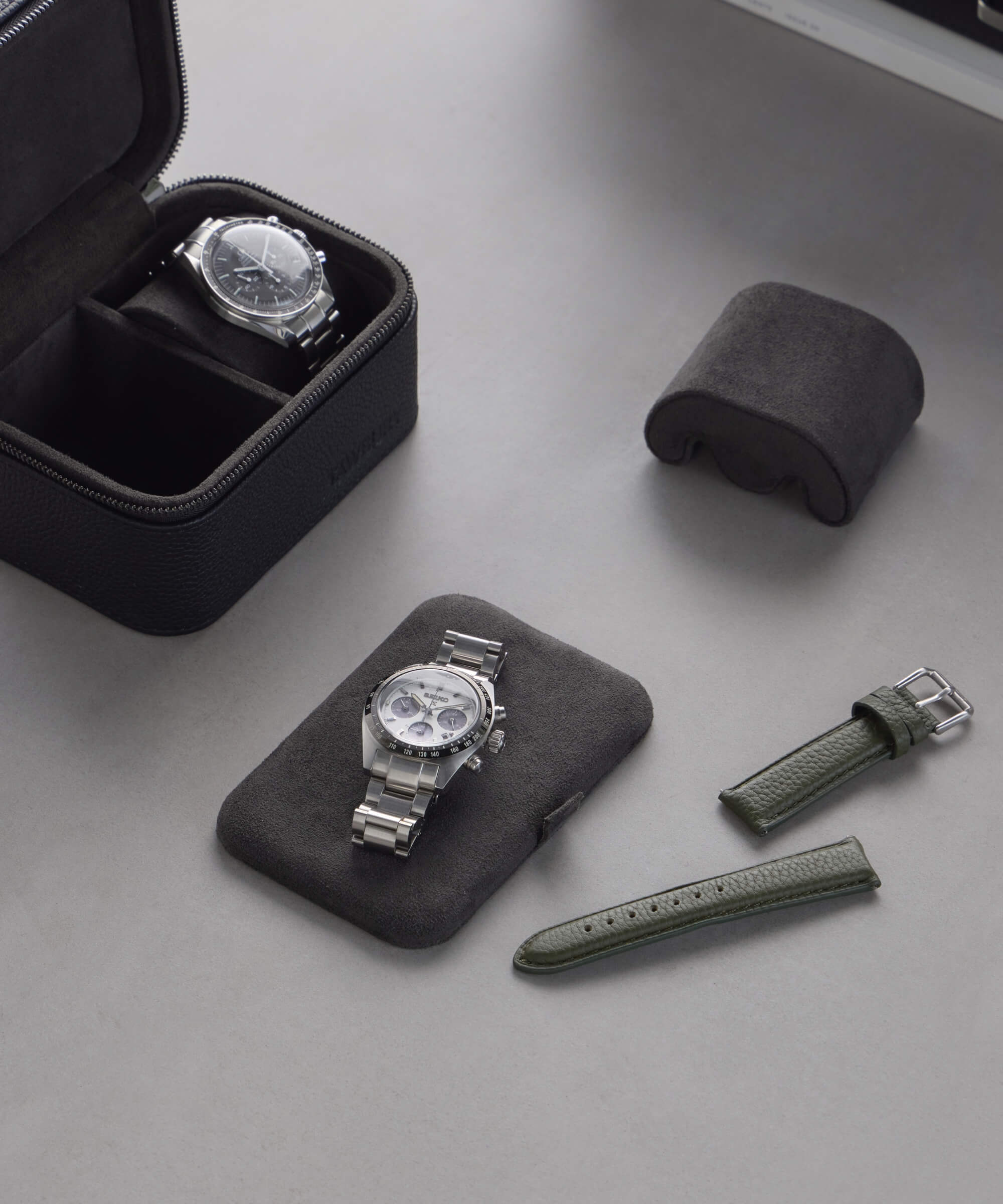 Two wristwatches: one in a Fraser 2 Watch Travel Case - Black and the other elegantly displayed. With a green leather strap resting on a black cushion, these pieces from TAWBURY exude timeless style on the gray surface.