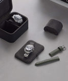 Two wristwatches: one in a Fraser 2 Watch Travel Case - Black and the other elegantly displayed. With a green leather strap resting on a black cushion, these pieces from TAWBURY exude timeless style on the gray surface.