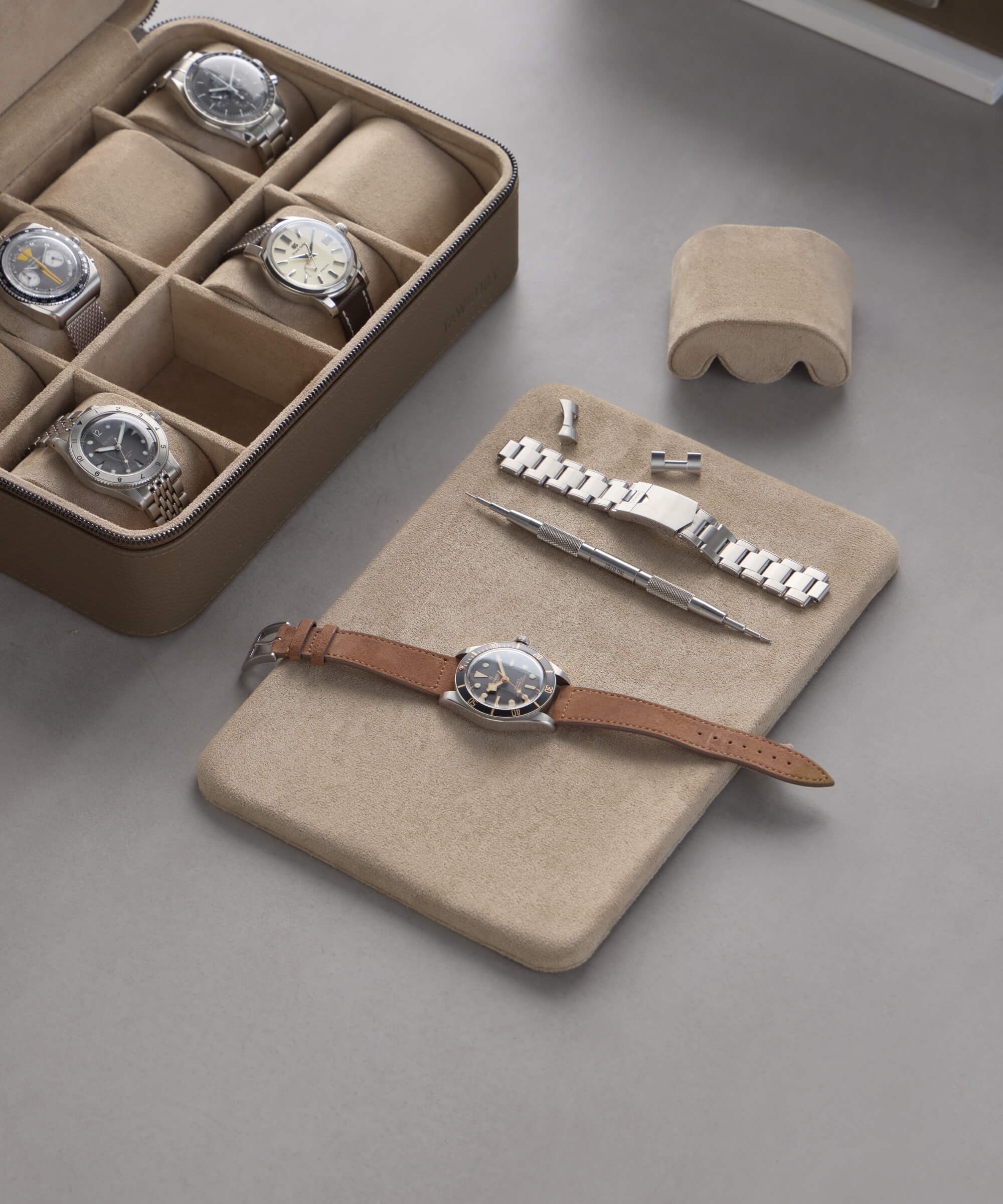 The TAWBURY Fraser 8 Watch Travel Case in taupe elegantly showcases a leather watch, tools, and a bracelet on a matching pad. This luxurious beige leather case sits gracefully on a gray surface, perfectly blending style and function.